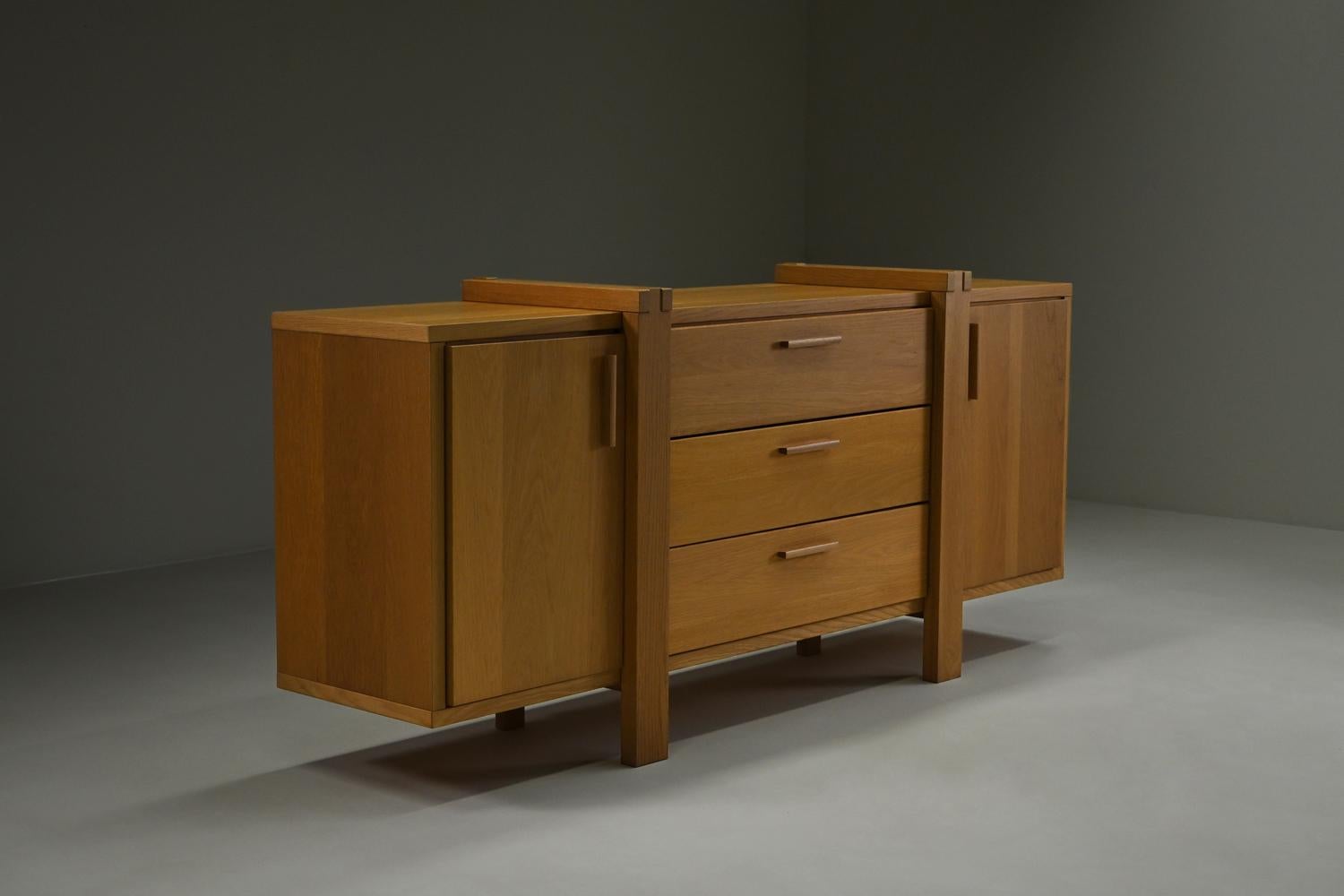 This Belgian sideboard is crafted mostly from solid oak and reflects the Brutalist style with its strong lines and architectural details. The piece offers plenty of storage with 2 cabinets and 3 drawers. Its natural oak finish brings warmth and