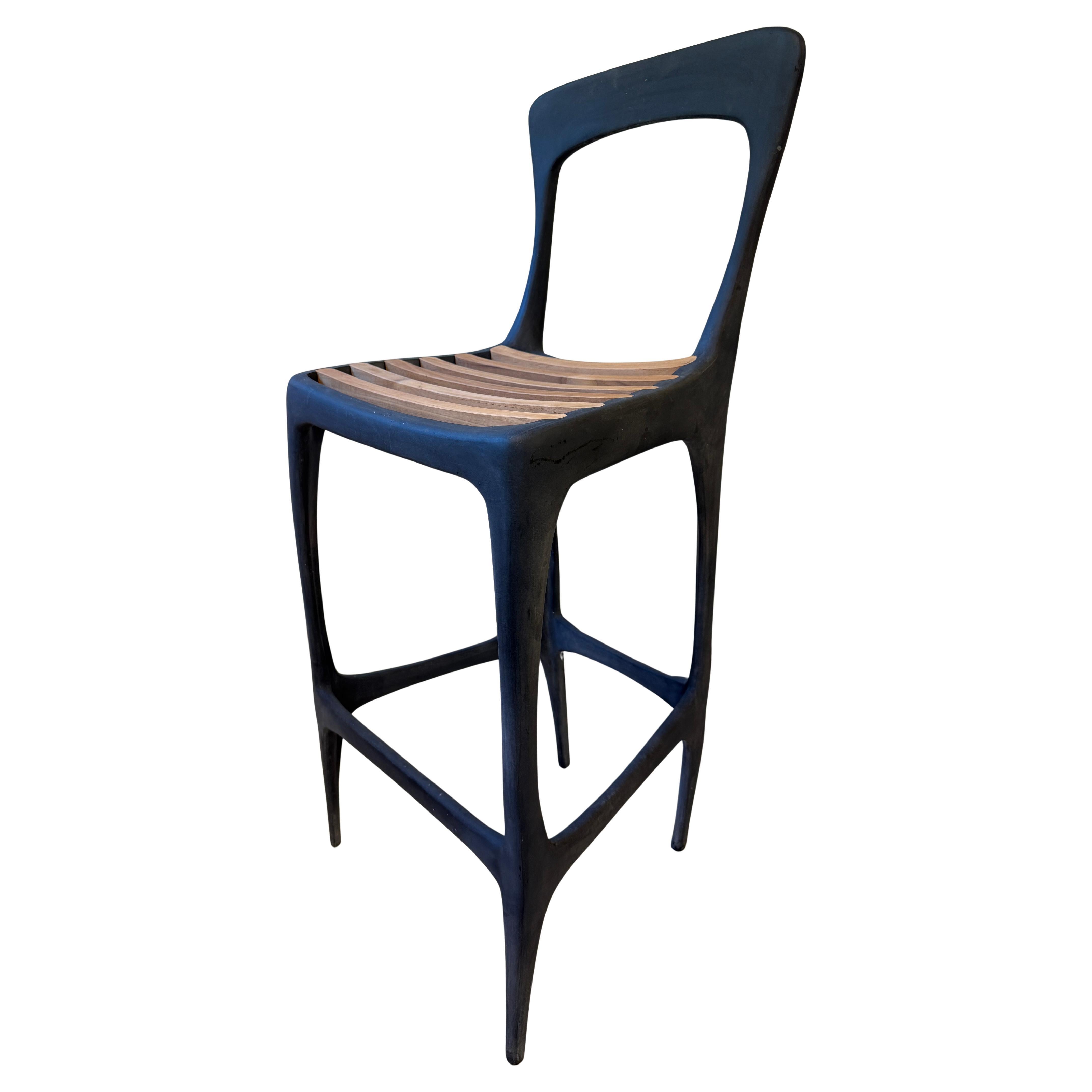A sculptural bar stool from the Flow Collection, designed by John Reeves for Henry Hall Designs, crafted from 100% recycled aluminum with a heavy zinc-plated finish and solid FSC-certified teak wood slats forming the seat. With its organic brutalist