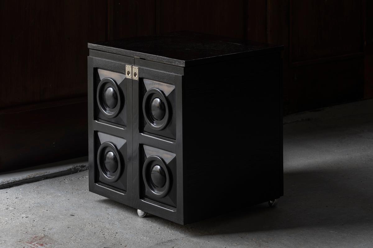Bar cabinet attributed to De Coene, Belgium, 1970’s. This black lacquered cabinet offers you space to store your liquor.  From compact to wide with an easy movement. The graphic door panels ensure a brutalist twist and the 3 wheels over you an easy