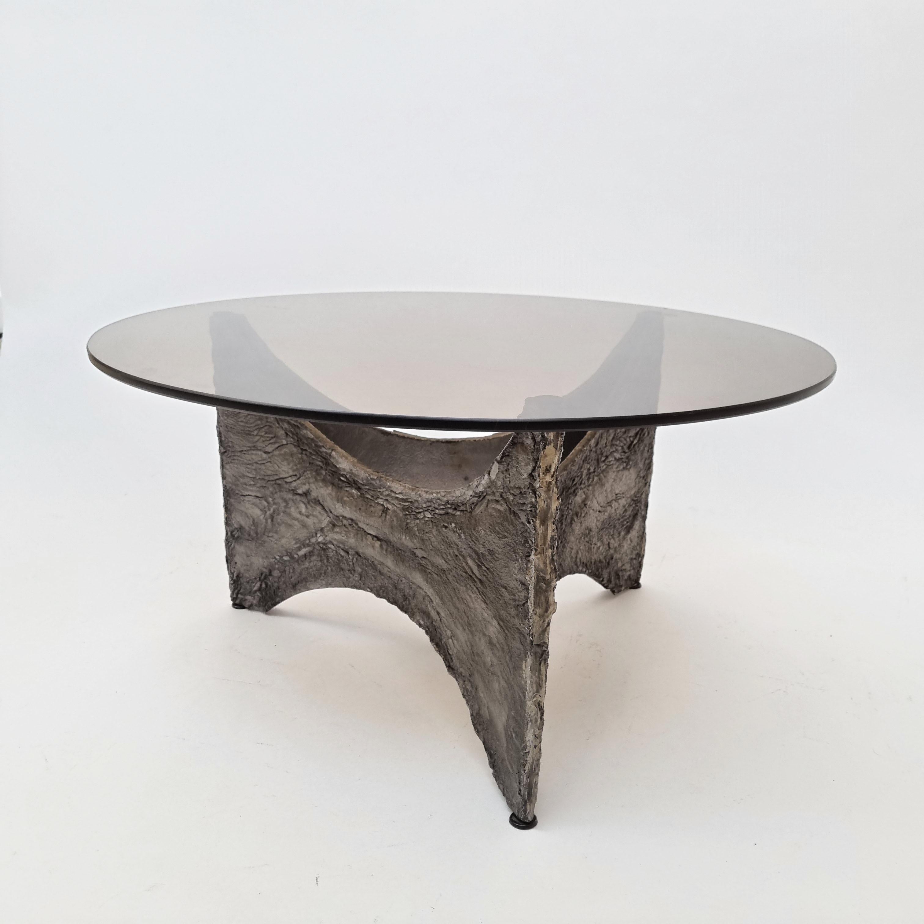This Brutalist coffee table is made in a style reminiscent of designs by Paul Evans. It was manufactured in Belgium, circa 1960. The table base is composed of cast aluminum with a rough texture. The top is made of smoke glass, revealing the shape