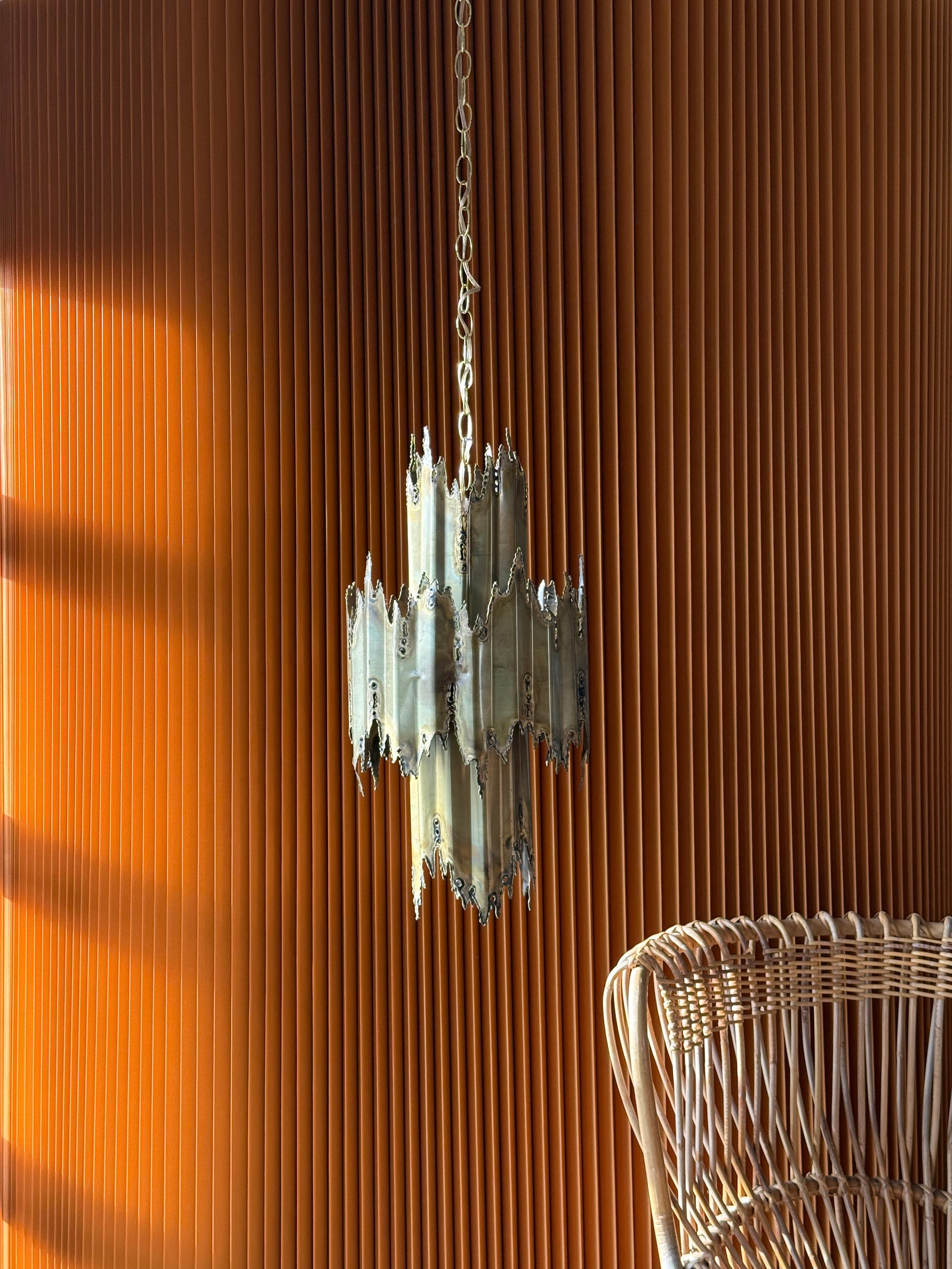 Magnifique lustre brutaliste du milieu du siècle, réalisé par le célèbre designer Tom Greene pour Feldman Lighting Co. vers les années 1960-1970. Ce luminaire sculptural met en valeur la technique de laiton découpé au chalumeau, signature de Greene,