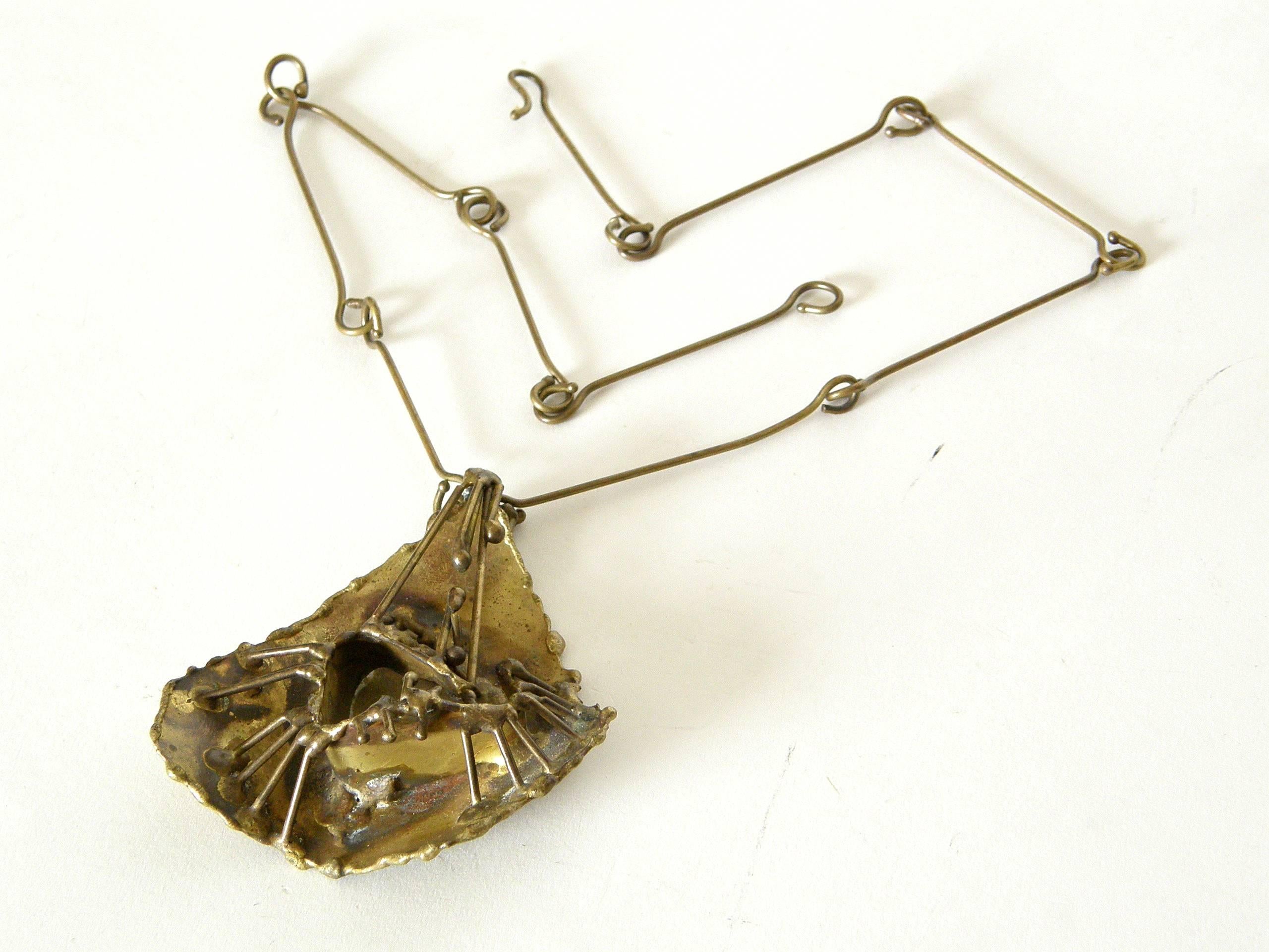This hand wrought brass necklace features an abstract pendant constructed in fairly high relief for the scale. It has torch cut edges, accents of oxidation, and a kind of architectural, weblike structure. It hangs from a chain of long, wire links.