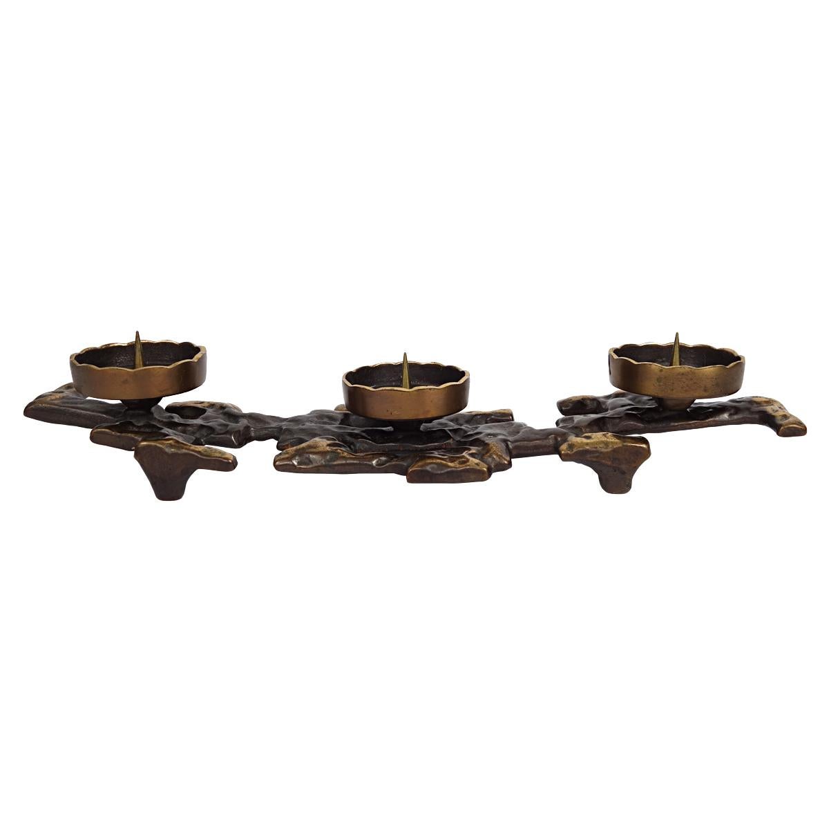 Stylish bronze candlestick of the rough Brutalist kind. 
It almost seems as if the molten bronze did not go into a mold but found its own way to shape up into an object. 
Four elegant feet raise the candlestick gracefully from the surface. 
Three