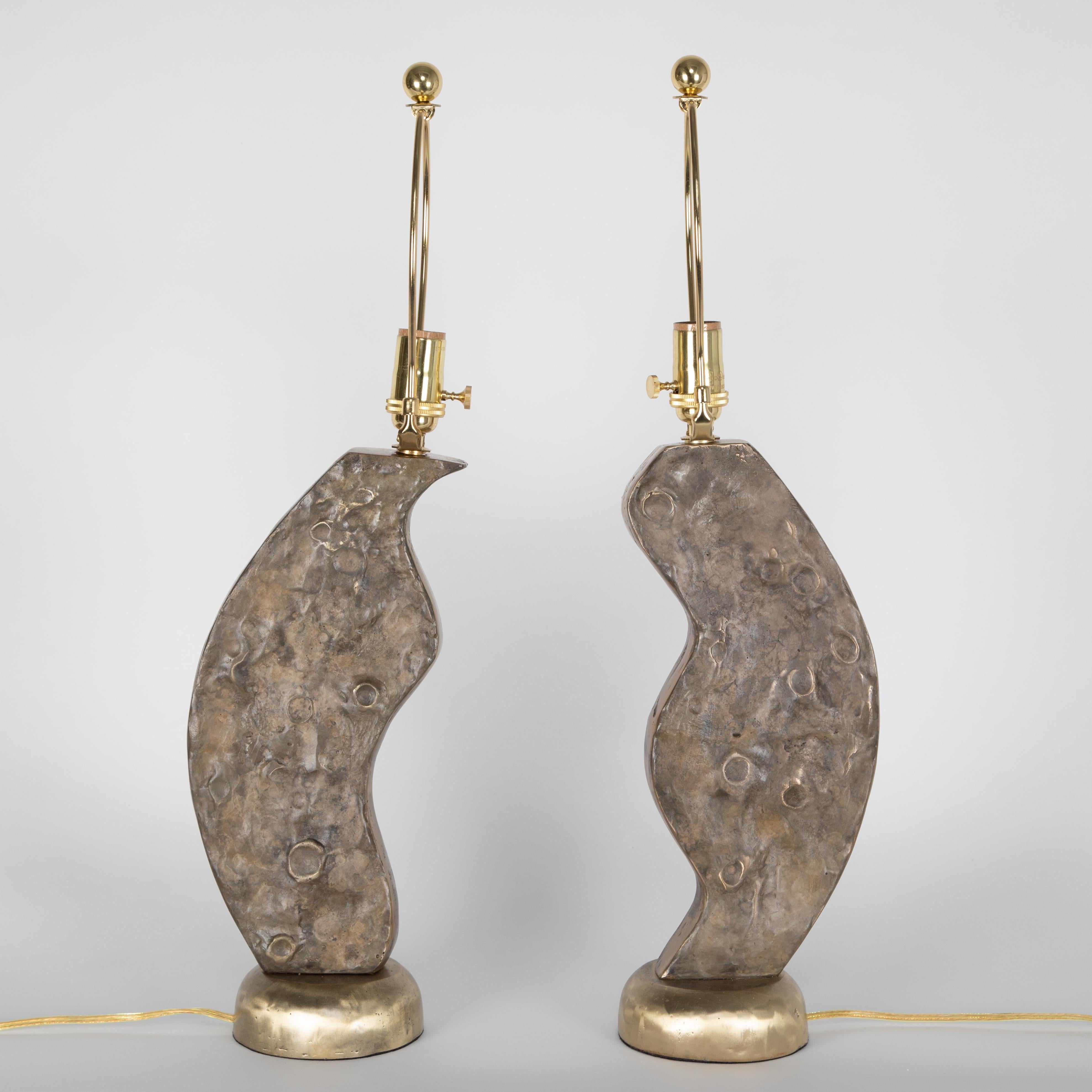 Pair of heavy Brutalist brass table lamps in complementing forms that would interlock like a puzzle if connected together. Fronts and backs are textured with crater-like details, and the sides and circular bases feature a shinier finish.