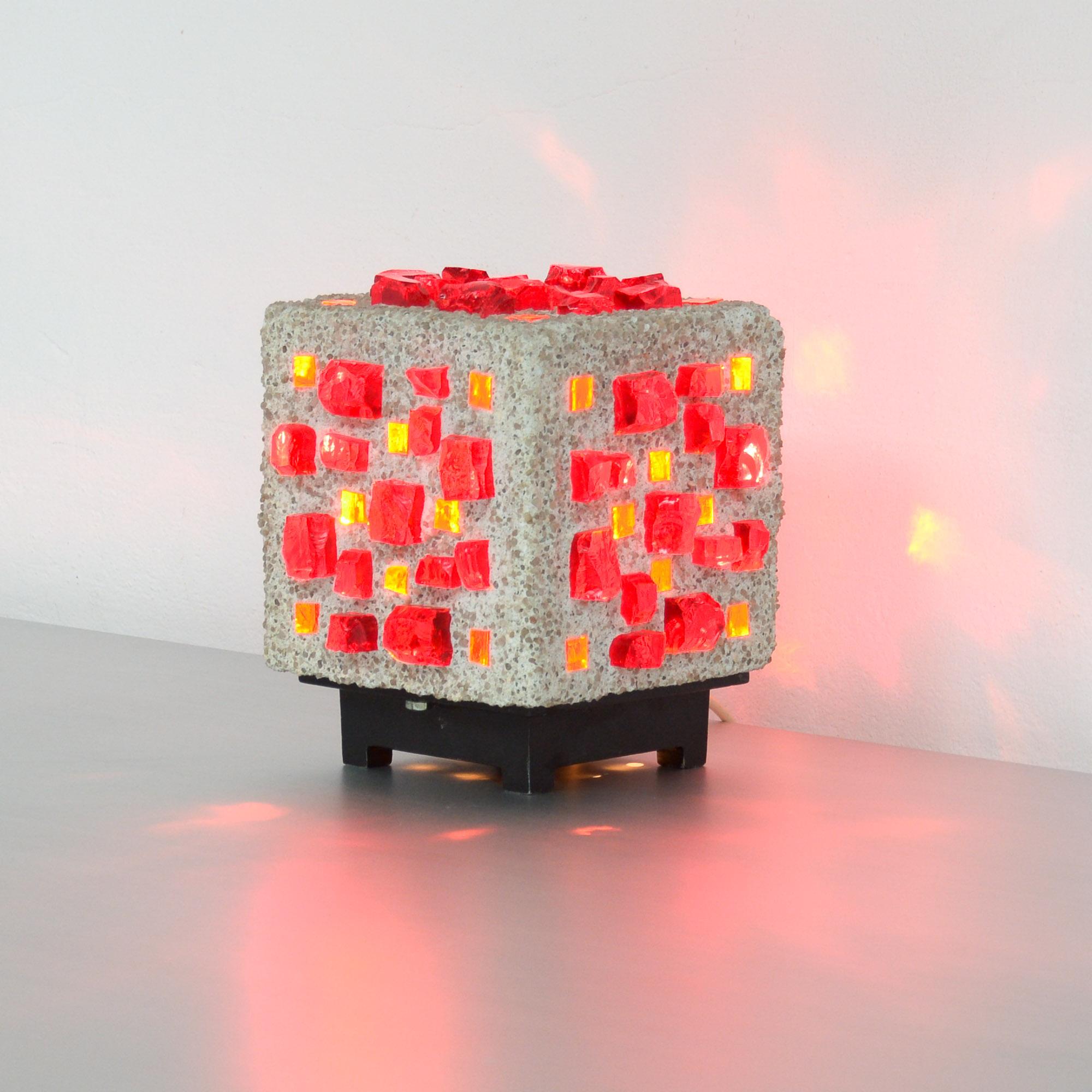 This Brutalist cube table lamp can be dated in the 1960s.
It is made of concrete and thick broken glass pieces in different shades of red.
The lamp was inspired by the Brutalist architecture and the stained glass windows of Le Corbusier.
It is a