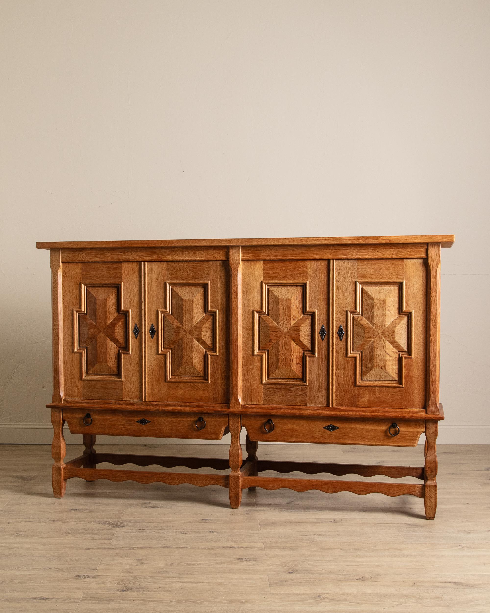 This stately vintage cabinet exudes craftsmanship and old-world refinement. Constructed from richly grained wood, it features four paneled doors adorned with deeply carved geometric motifs that create a striking play of depth and shadow. Resting on