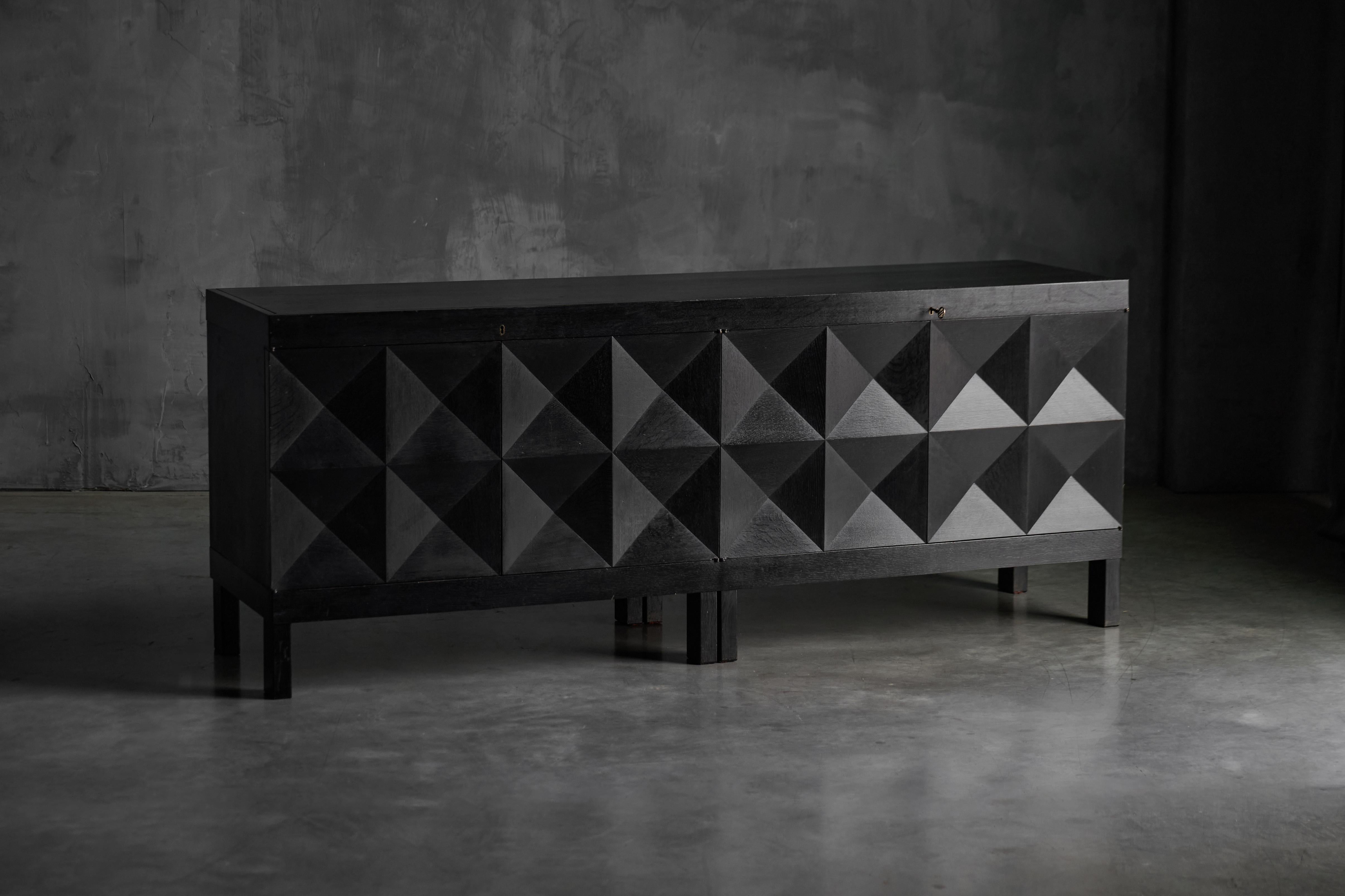 A brutalist sideboard by De Coene, made in Belgium in the 1970s. Crafted from black-stained oak, the piece showcases four geometric diamond-pattern doors opening to reveal generous storage. Its rhythmic symmetry and strong graphic presence define