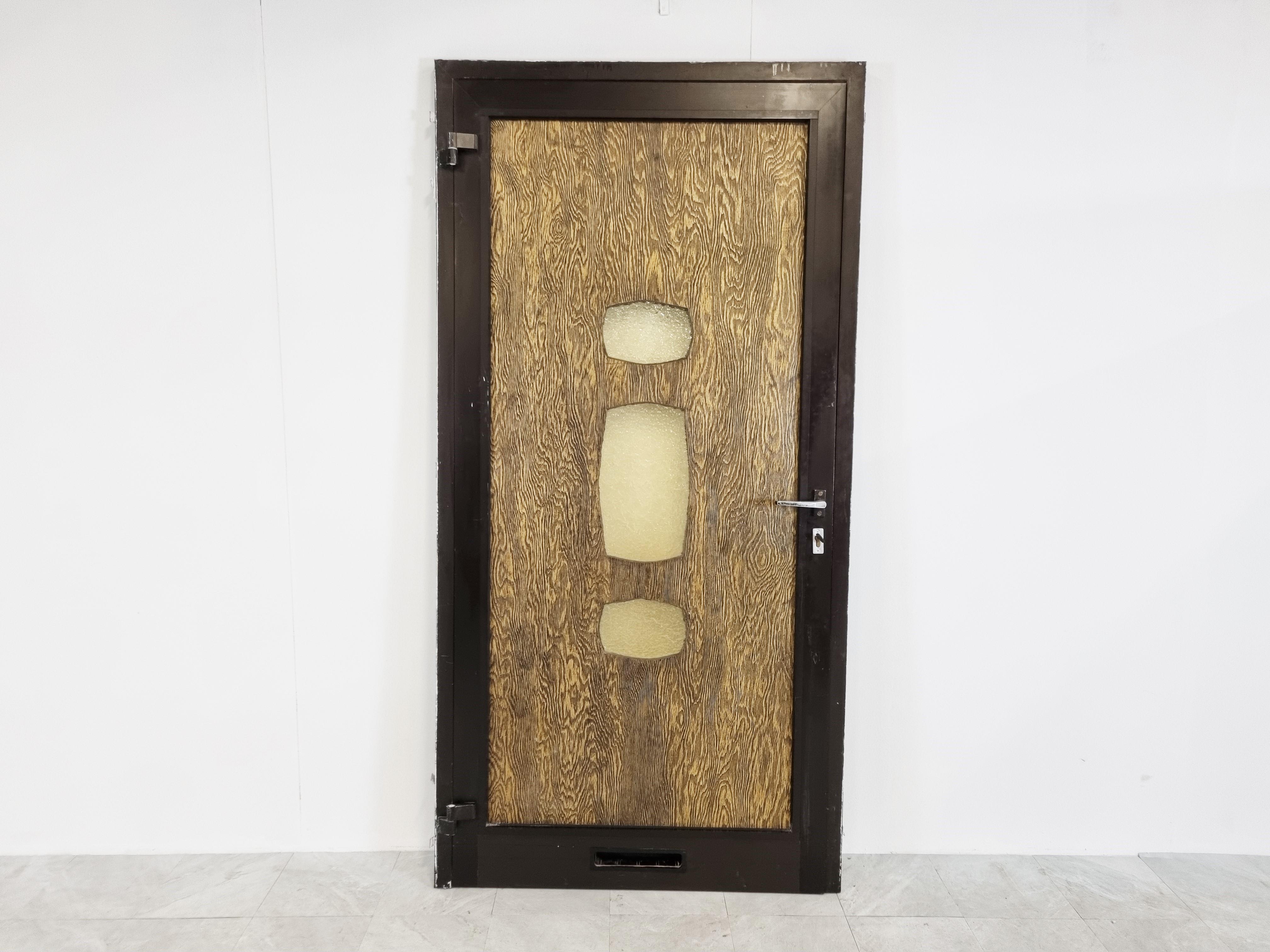 Architectural brutalist front door with copper sculpture at the front, blurred glass windows and a wooden panel at the back.

This is a complete door with door frame.

1970s - Belgium

Measures: Height: 218cm/85.82