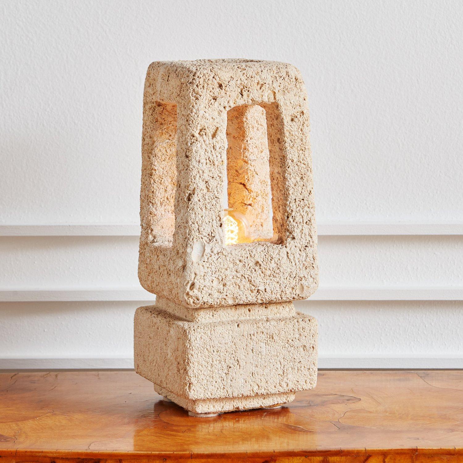 A brutalist French hand carved porous limestone lamp attributed to Albert Tormos. This sculptural lamp has a square plinth base and accepts one standard lightbulb which is visible through rectangular cutouts on all four sides of the lamp. There is