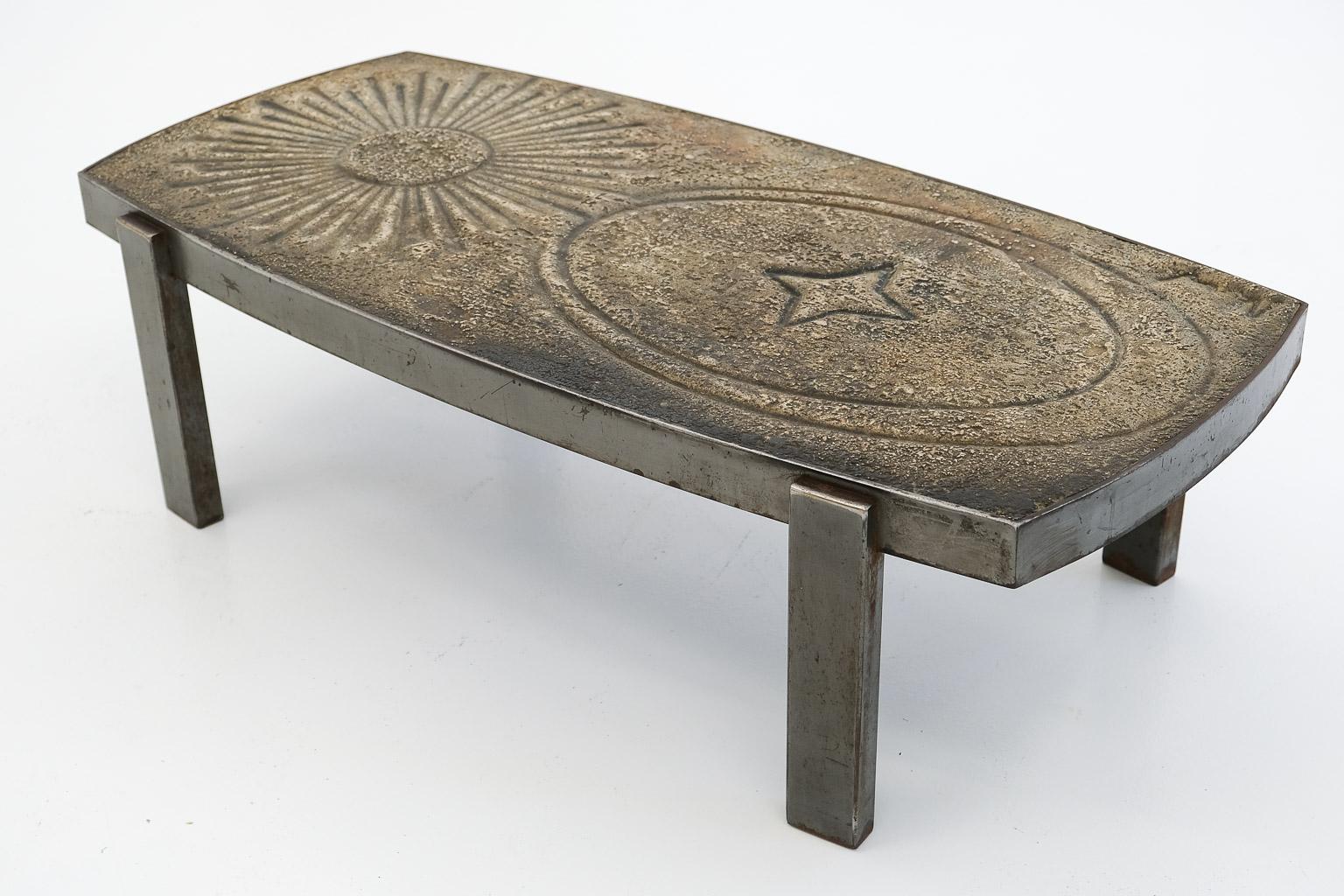 A heavy lava stone slab rests on an anthratzite-coloured steel frame. The lava slab shows a deeply embossed graphic form, a sun with several rays and a rhomb surrounded by ellipses. 

This table was designed and produced by the French ceramic artist