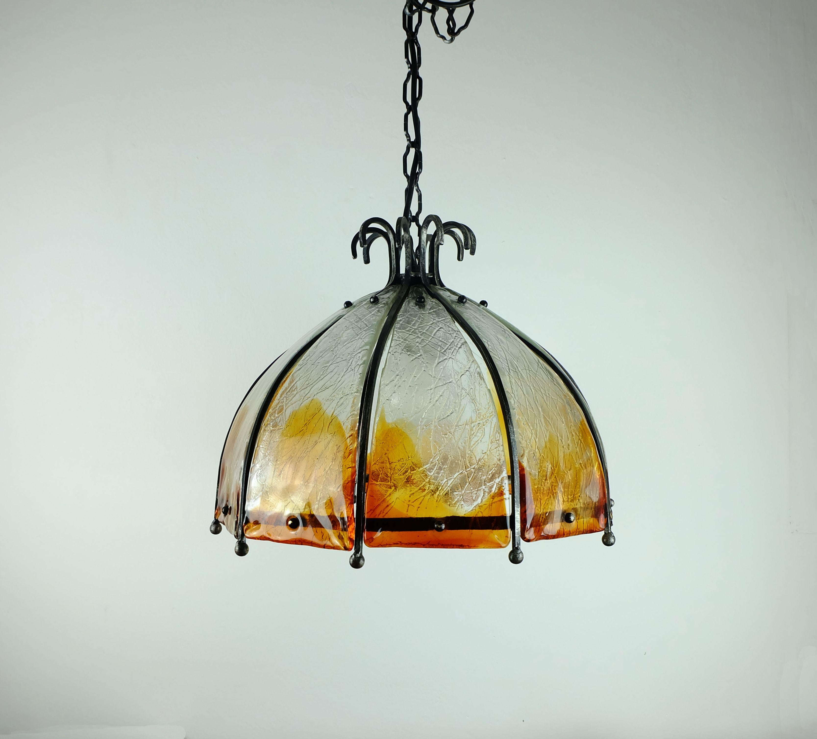 Una fantastica lampada a sospensione vintage italiana degli anni '60-'70. La struttura, la catena e il baldacchino sono in ferro battuto, mentre il paralume è in vetro spesso, trasparente e color ambra.

Il diametro è di 47 cm (18 1/2