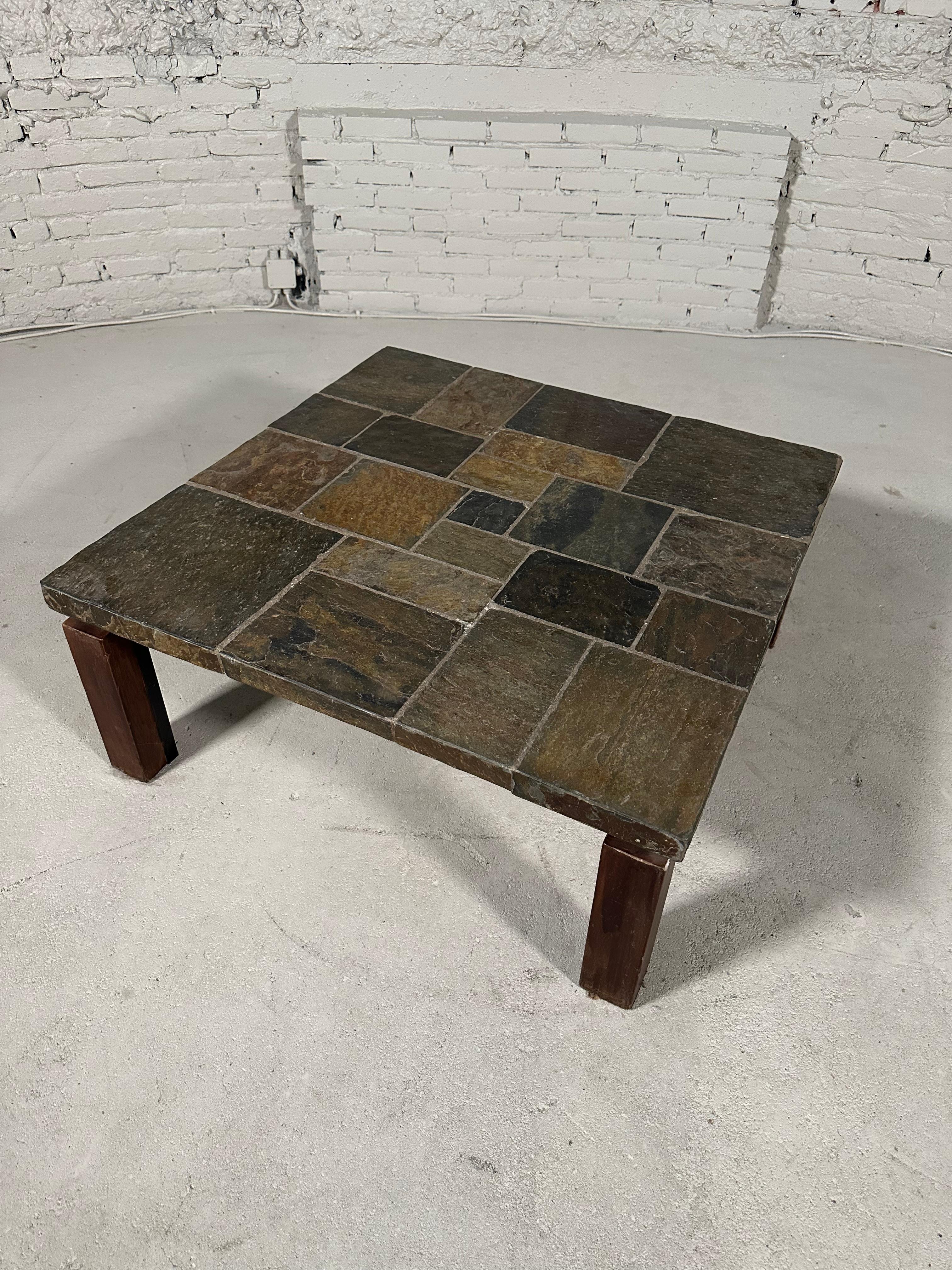 A square coffee table reminiscent of Paul Kingma's work, presumably made in the 1970's. 

The tabletop is constructed with squares of different types of natural stone. It rests on a black lacquered steel base with wooden legs. Wear is consistent