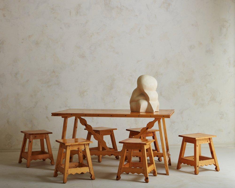 A brutalist 1950s Italian table constructed with pine wood. This table has four angled legs and features a sculptural stretcher with wood joinery and a carved cutout design. It has five matching pine wood stools with square seats and matching carved