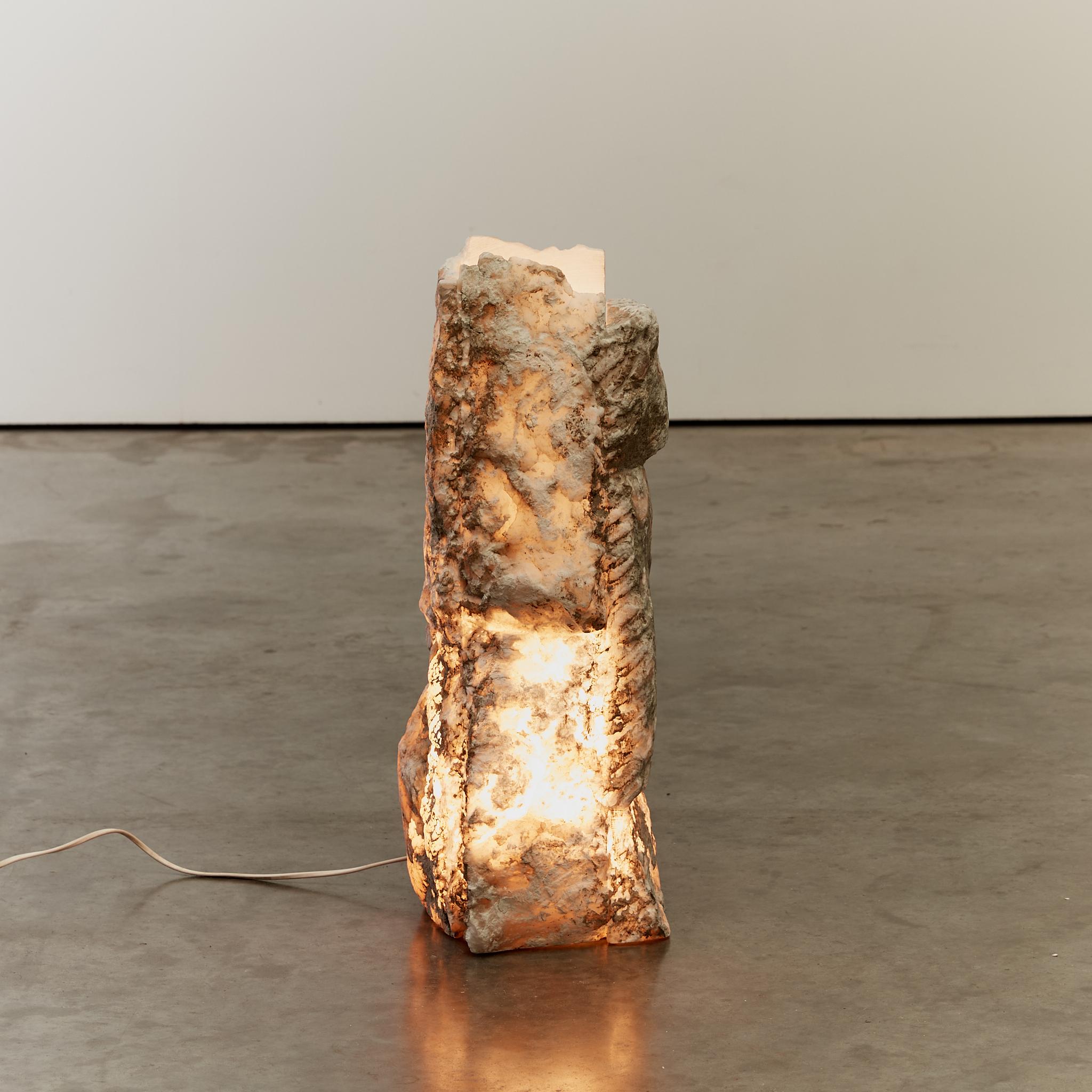 Raw finish column form lamp. Made from alabaster slabs, the thick stone allows for a subtle ambient light when lit. Measuring half a meter tall, this impressive lamp is either suitable as a table top piece or suitable for being situated on the
