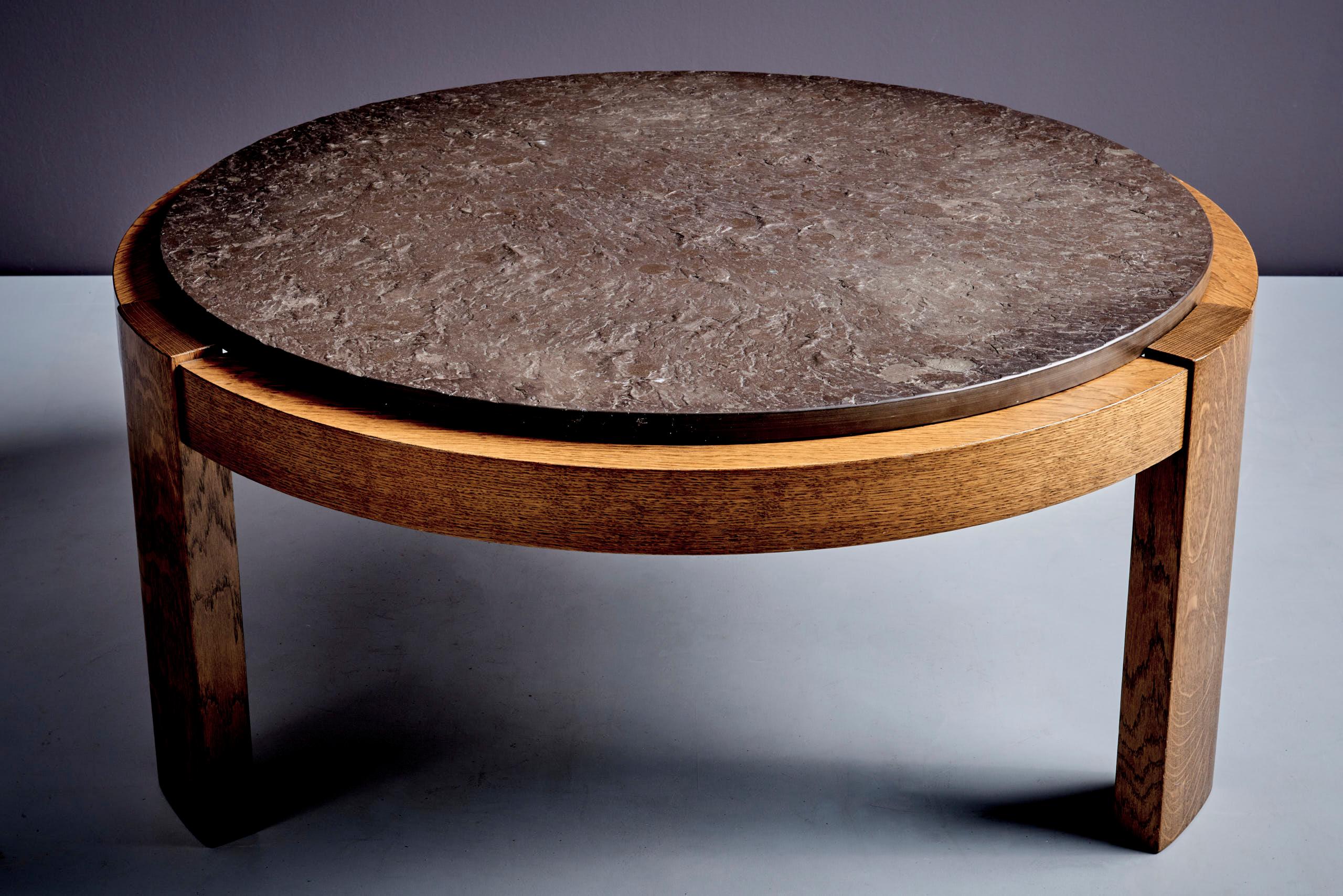 Table ronde brutaliste à trois pieds avec un plateau en pierre fossile foncée et un cadre en chêne. La partie supérieure présente une dalle de pierre fossile texturée, qui met en valeur les fossiles visibles incrustés dans la pierre. Elle est