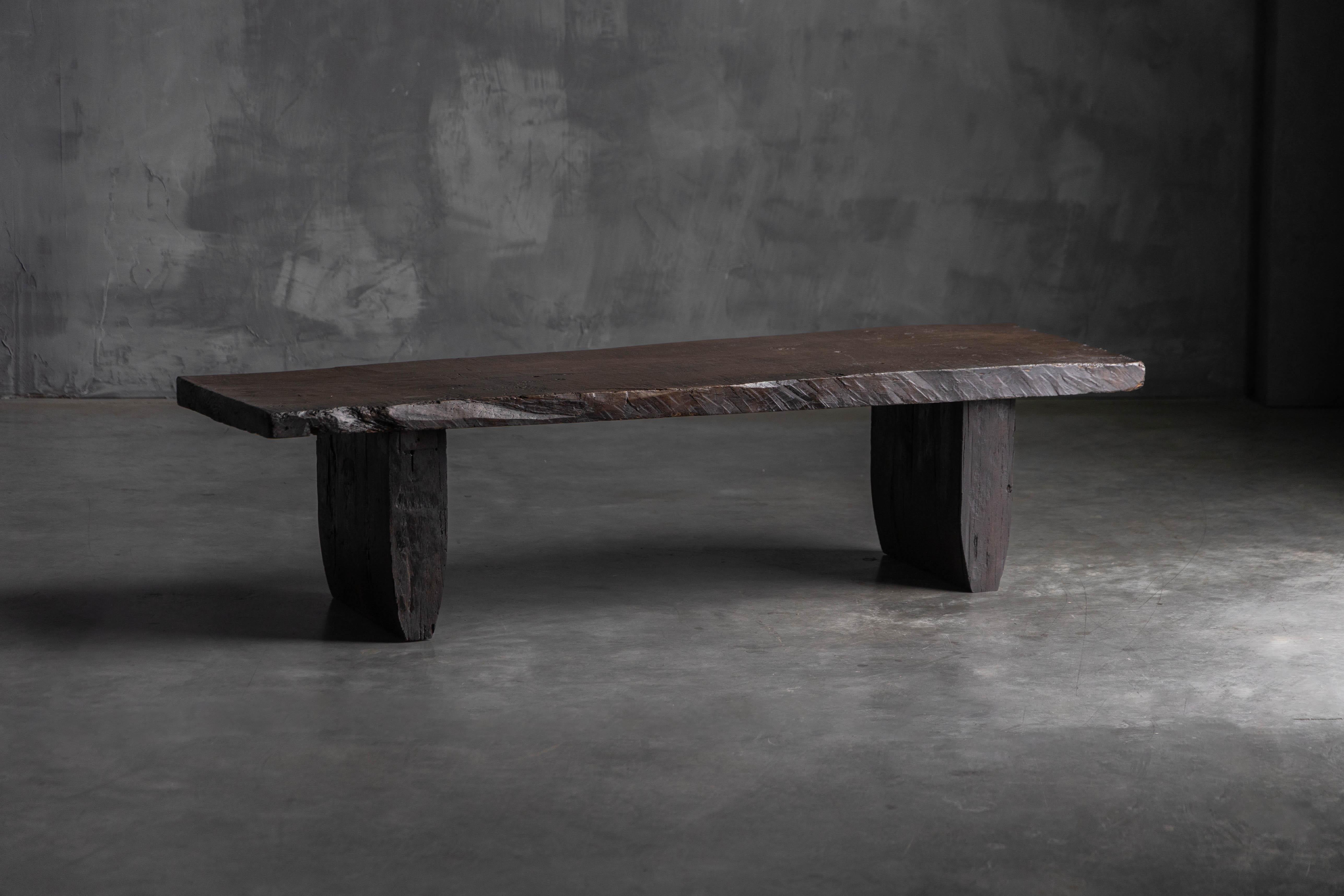 This late 18th-century rustic coffee table is crafted from a solid slab of dark, timeworn wood with a deep, natural patina. Its raw, irregular edges and visible tool marks speak to primitive craftsmanship, while the robust block legs lend it a