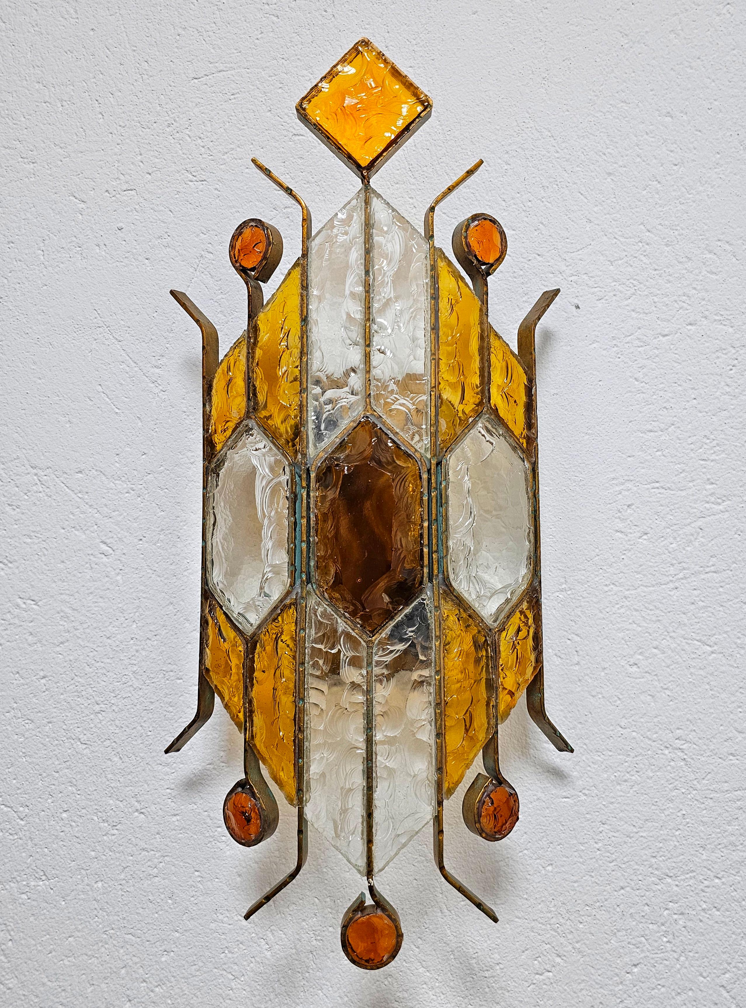 In this listing you will find a spectacular and large Brutalist sconce designed by Biancardi and Jordan Arte,  the main competitor of Longobard and Poliarte in Italy in the 1970s. It features a large fixture with hammered glass in amber and clear