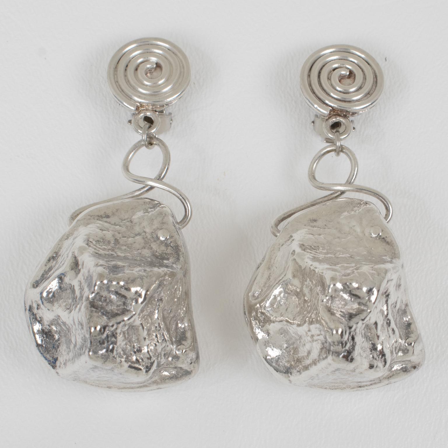 Ces superbes boucles d'oreilles pendantes à clip présentent un design brutaliste avec une goutte surdimensionnée en forme de pépite de roche en métal argenté. Les éléments de fixation présentent un motif en spirale en métal argenté. Il n'y a pas de