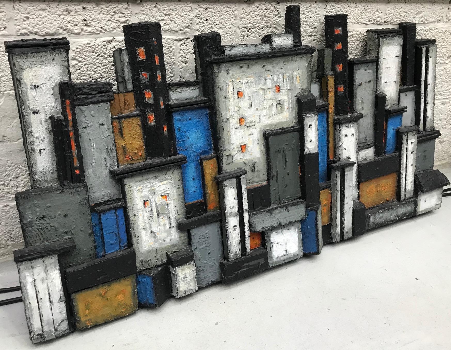 Skyline cityscape wall relief build in blocks of rough textured wood and painted in black, white, orange & blue is made from the perspective of the waterfront river side. It is made and signed by the Scandinavian artist Dorrit Kahler in the