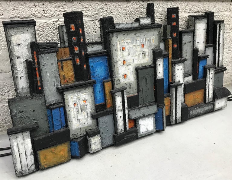 Skyline cityscape wall relief build in blocks of rough textured wood and painted in black, white, orange & blue is made from the perspective of the waterfront river side. It is made and signed by the Scandinavian artist Dorrit Kahler in the