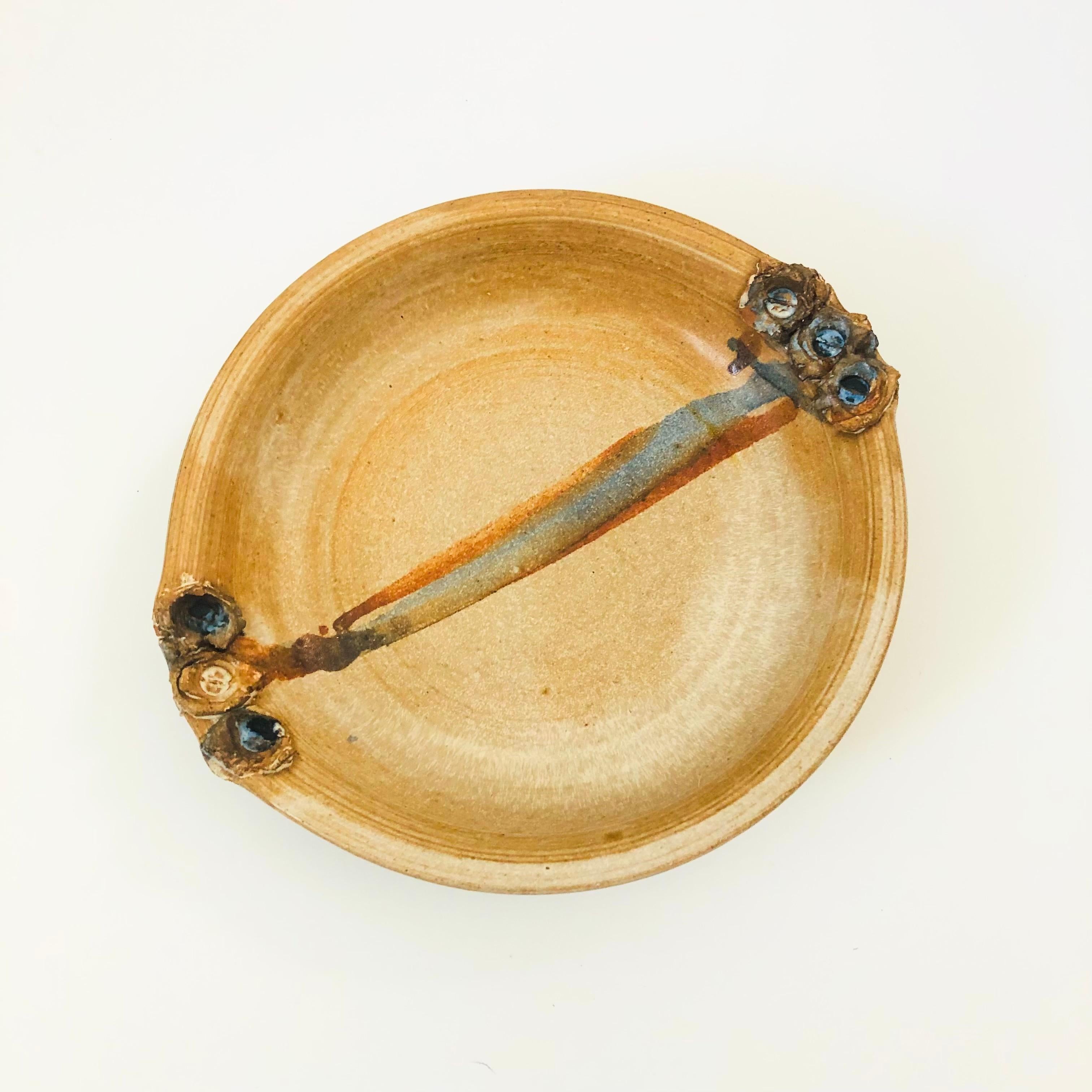A vintage circular studio pottery tray. Earthy glazes with unique textured handles on the sides. A stripe of contrasting blue and brown glazes connects the 2 handles. Embossed signature of the maker on the side.

