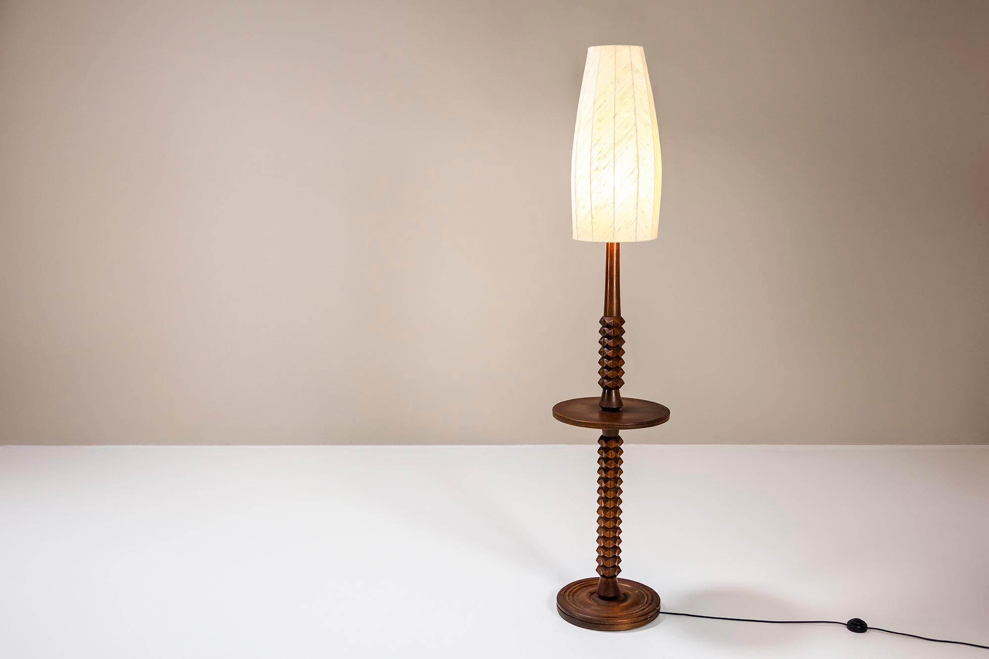 Esta lámpara de pie presenta un diseño limpio y funcional con una estructura de madera de roble en acabado marrón silla de montar. La lámpara alta está rematada con una pantalla de tela beige que difunde la luz suavemente, creando un cálido