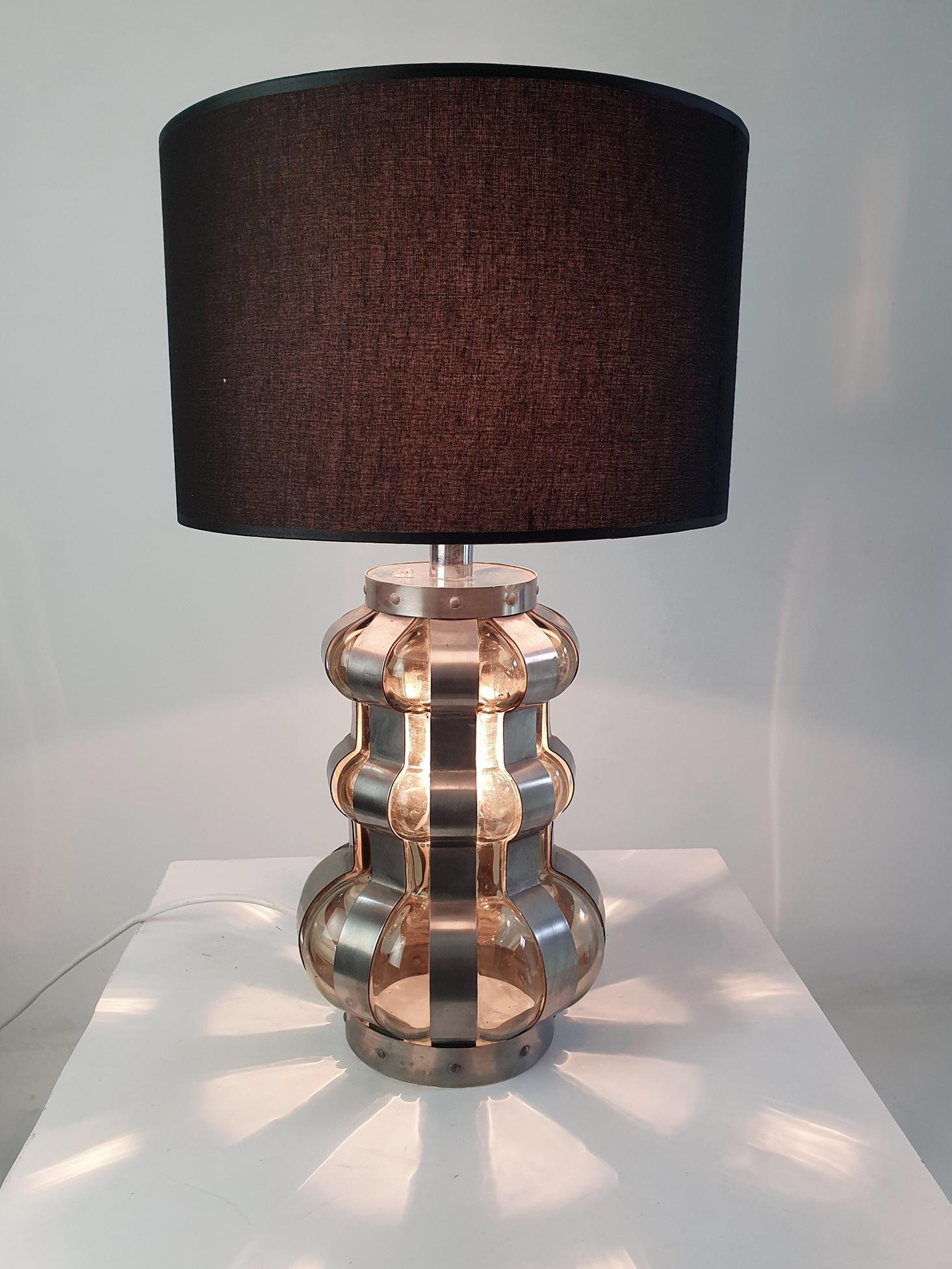 A large table lamp from the 1960s with a base in smoke colored glass which is lit from the inside and caged in by a metal chrome construction. The diameter of the base of the lamp is 22 cm and the lampshade is 35 cm in diameter. Total height with