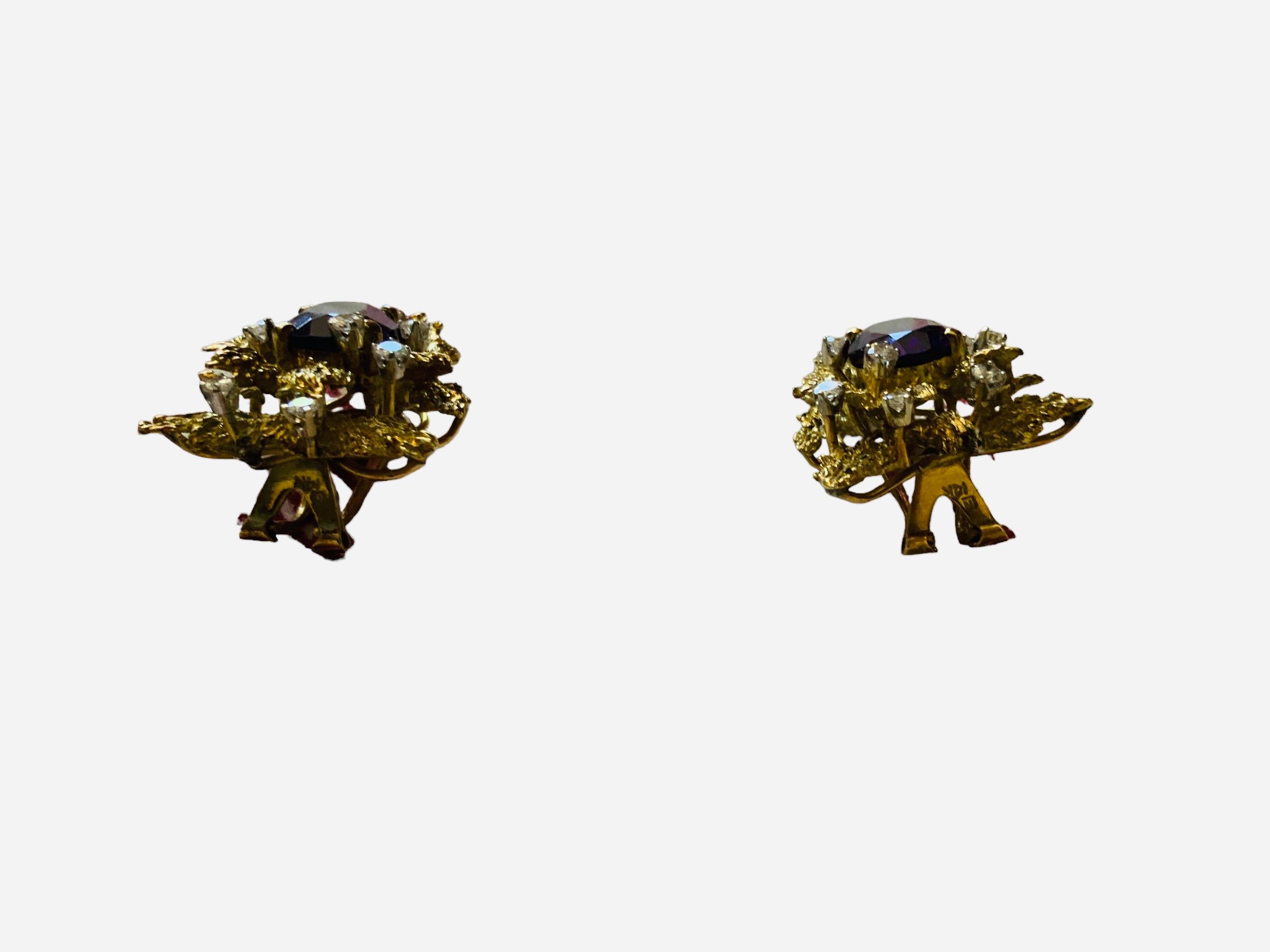 This is a Brutalist style pair of 14K yellow gold, Amethyst and diamonds omega clip earrings. It depicts a pair of oval faceted amethysts mounted in prong setting. They are decorated with 14K gold “wood and /or rock shaped pieces” and enhanced with