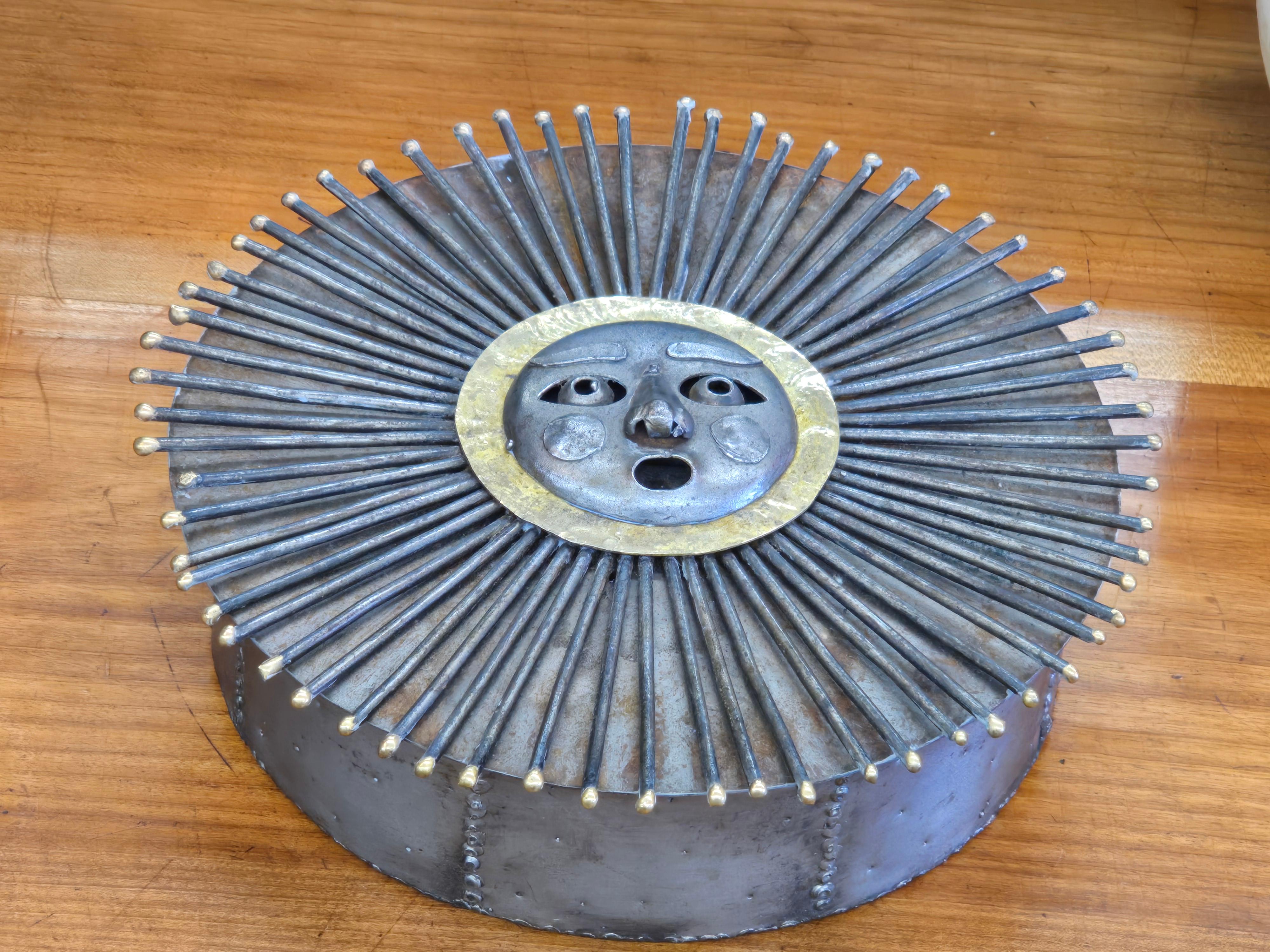 Eine handgeschmiedete brutalistische Metallbox in Form einer strahlenden Sonne, hergestellt von den Emaus-Werkstätten in Mexiko. Der Deckel zeigt ein stilisiertes Gesicht mit ausdrucksstarken Zügen, umgeben von vergoldeten Akzenten und