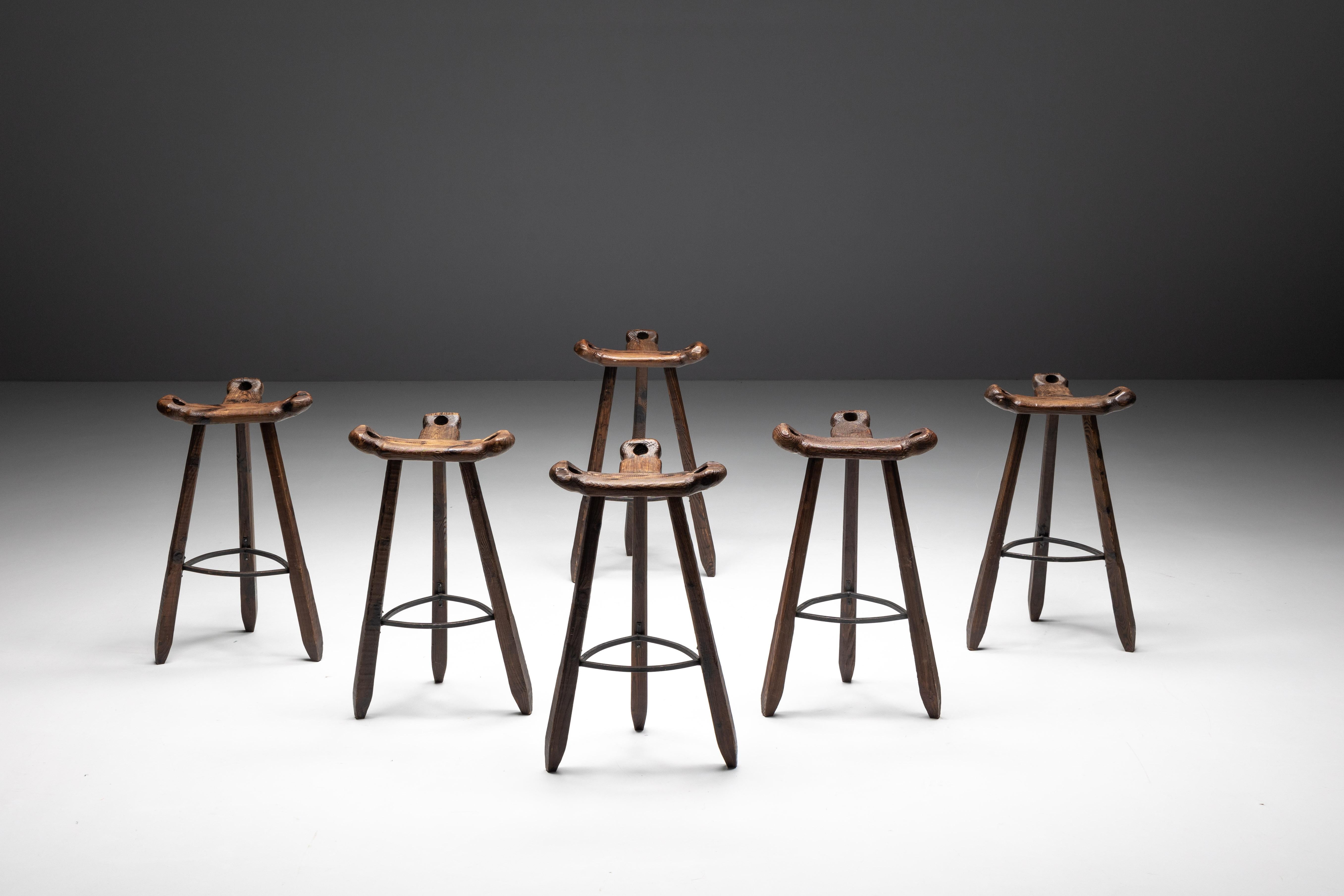 Spanish oak tripod bar stools designed with the distinctive influence of Brazilian designer Sergio Rodrigues. These stools feature ergonomic curved T-shaped seats with handles on all sides and three sturdy tapered legs equipped with a forged steel