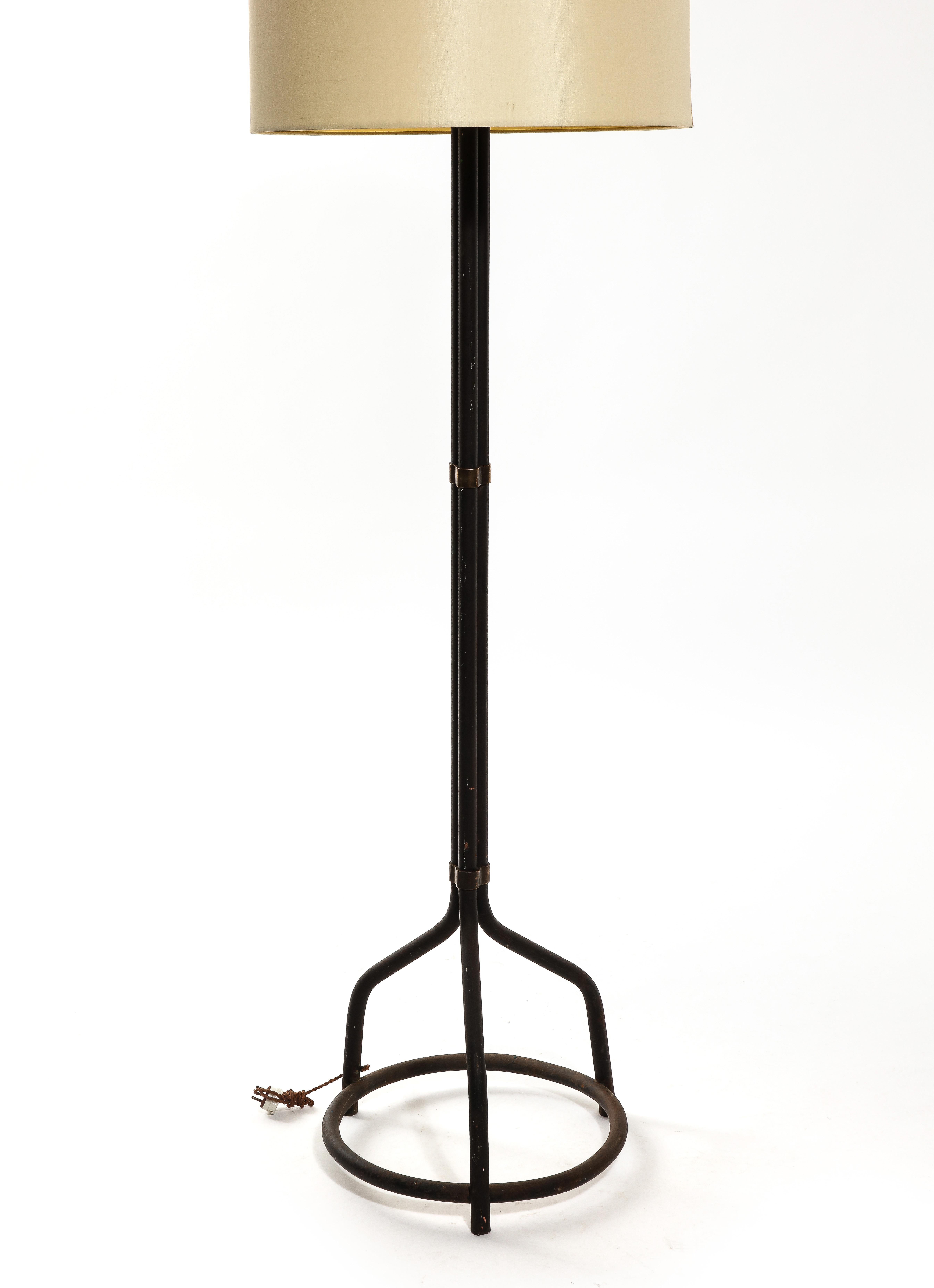 Industrial brutalist floor tripod flloor lamp made of black painted steel pipes with clover shpaed brass links on lower, mid and top sections. The tripod feet structure is conected by a circle horizontal bar which contrasts with the general graphic