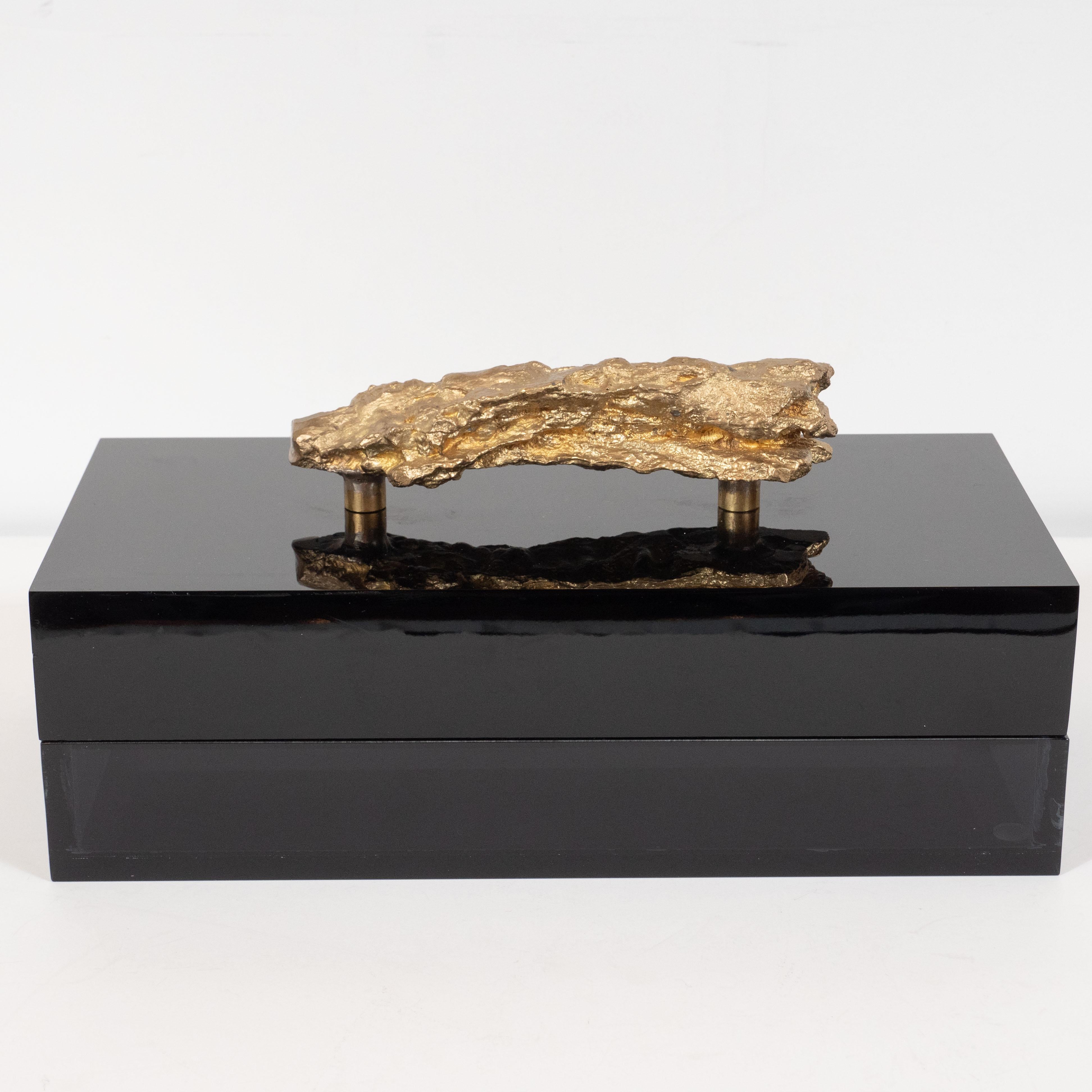 This sophisticated modernist Brutalist box offers a volumetric rectangular black Lucite body with an organic abstracted gilded metal pull suggesting an oversized gold nugget or a large fossilized stone. The organic texture of the cast metal stone