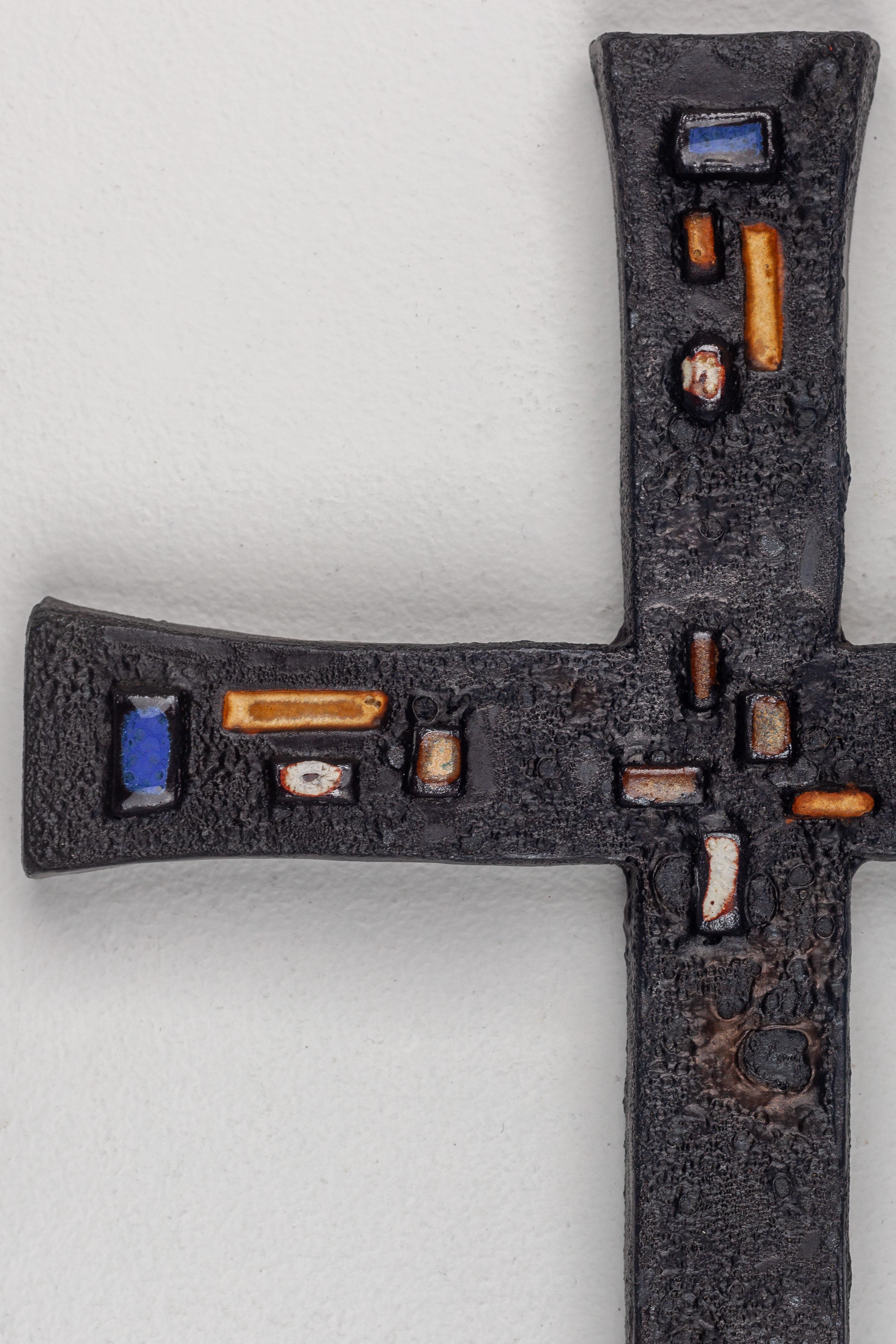 This exceptional Brutalist wall cross features a distinctive texture reminiscent of lava on rough stone, with subtle hues of blacks, sometimes exhibiting metallic accents. The charismatic black cross is adorned with long, thin, glossy glazed color