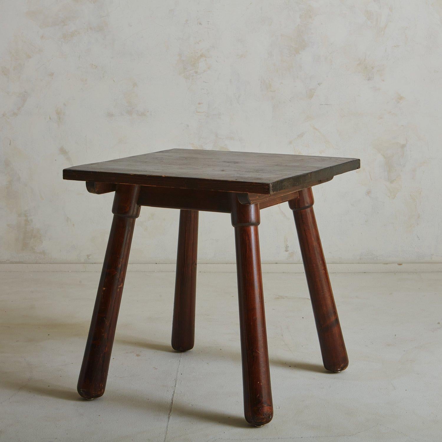 A brutalist 1960s French wooden dining table attributed to Georges Robert. This table has a rich walnut stain and a sculptural profile. It has a square top and stands on four angled, cylindrical legs with rounded feet. Unmarked. Sourced in France,