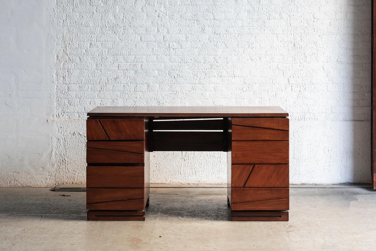 Writing desk designed and produced in the 1980’s by an unknown maker. The geometrical lines on both sides of the desk make this one stand out. They hide 3 drawers on the left and a cabinet with simple shelf on the right. The dark stained wood is in
