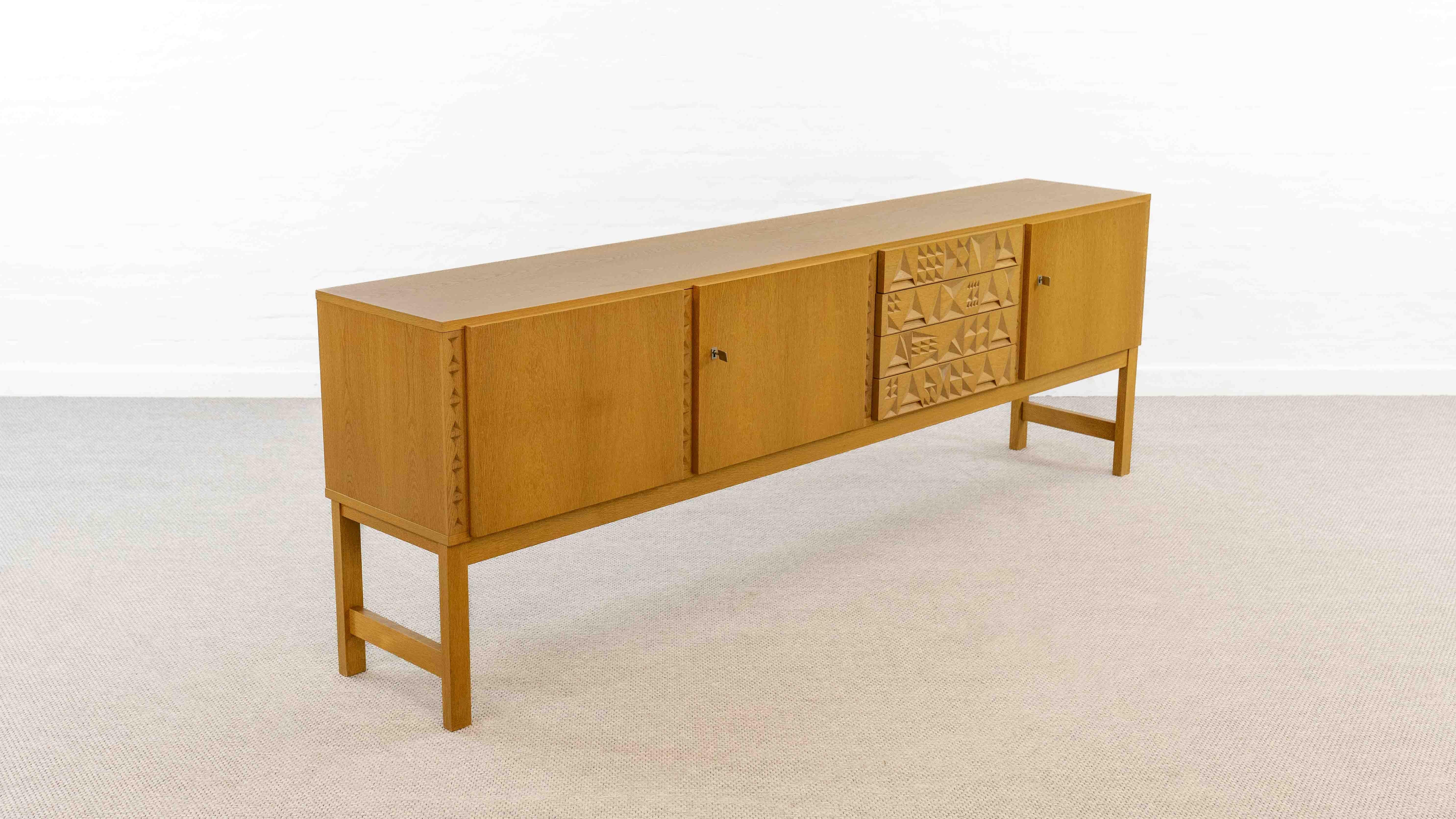 Credenza di metà secolo degli anni '60/'70. Realizzato in impiallacciatura di Oak. Frontale dei cassetti e cornice con ornamenti geometrici. Dispone di un ampio spazio di archiviazione con 3 porte e 4 cassetti. Due chiavi incluse. In buone