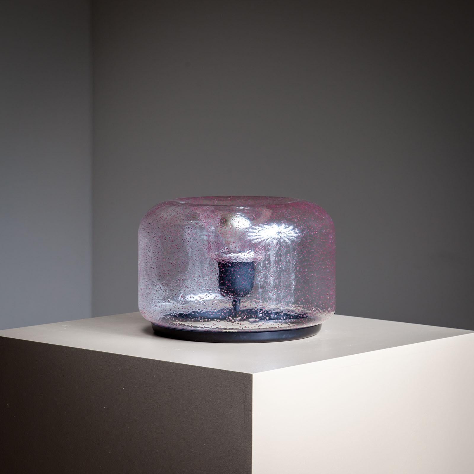 Lámpara de sobremesa redonda con pantalla anular de cristal gris con parpadeos rojos encerrados. La bombilla se sitúa en el centro y penetra en el cuerpo de cristal desde el interior. La etiqueta VeArt con el topónimo 