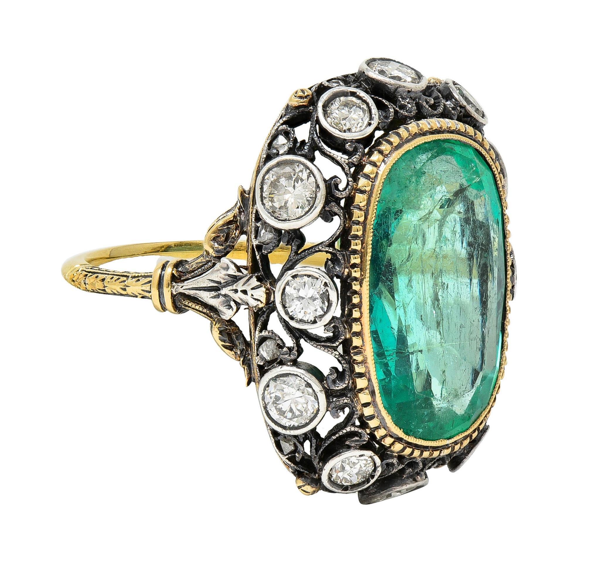 Centering an elongated cushion cut emerald weighing 7.10 carats total - transparent light green in color 
Natural Colombian in origin - set with a milgrain and groove detailed gold bezel 
With a domed sterling silver gallery surround with scroll