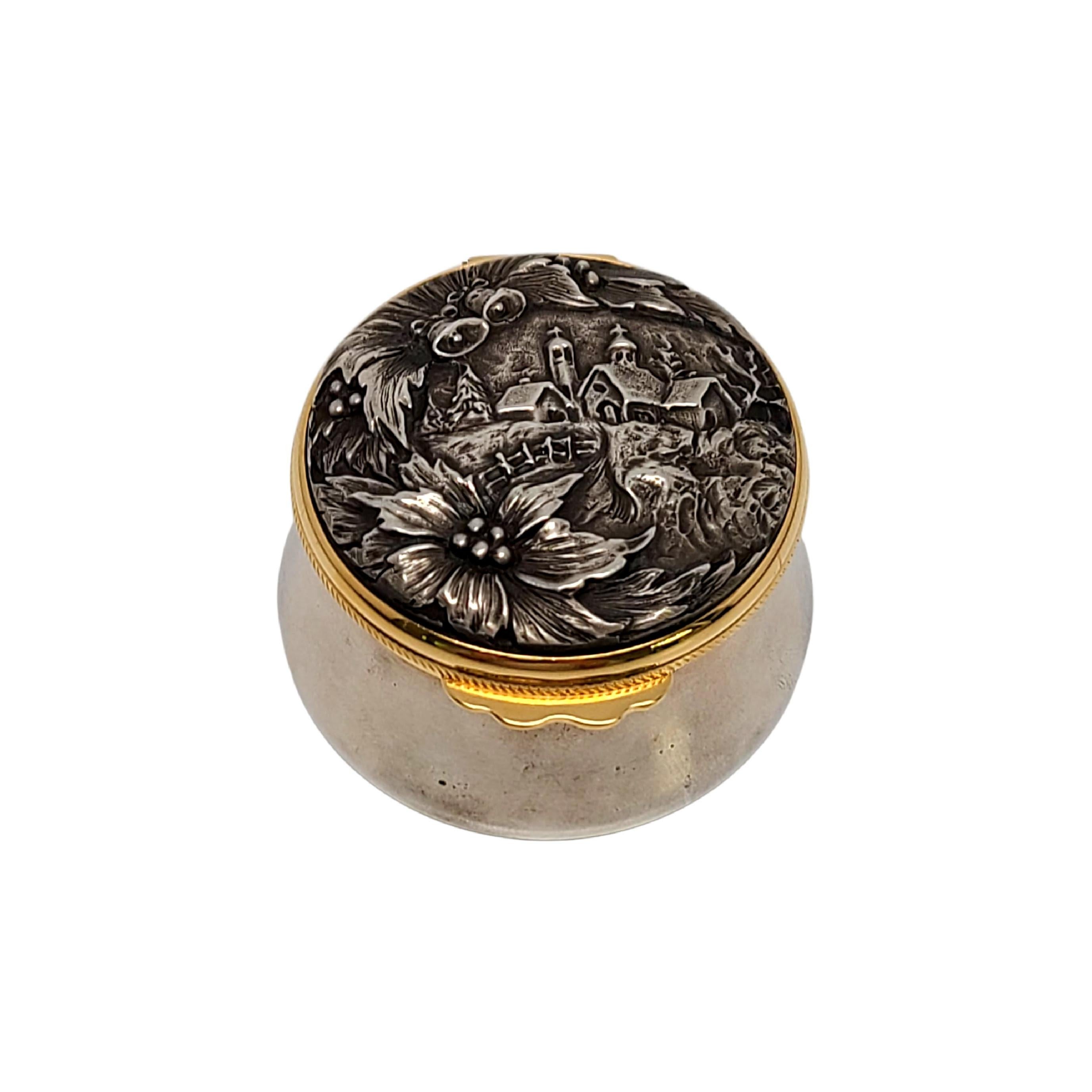 Round sterling silver box with gold rim by Buccellati.

This beautiful piece features a high relief Christmas scene on the lid, including a church, bells, mistletoe and holly.  The lid has a gold rim and a mirror inside. Designed by one of the most