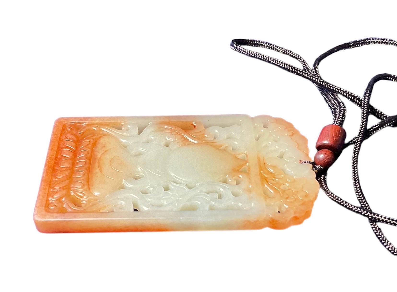 This a Buddha Jade Pierced Pendant. It is a rectangular hand carved white and orange jade depicting a seated Buddha and adorned with scrolls and flowers. The pendant is tied to a braided thread.