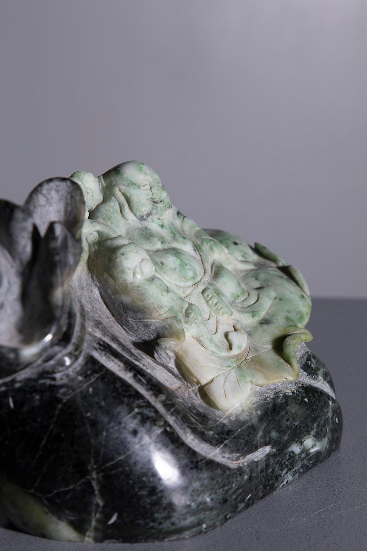 Statue en racine de jade vert et noir, représentant un Bouddha joyeux reposant sur un grand sac qui s'enroule gracieusement autour de lui. La figure est sculptée avec élégance et maîtrise : le Bouddha a une expression sereine et joyeuse, dégageant