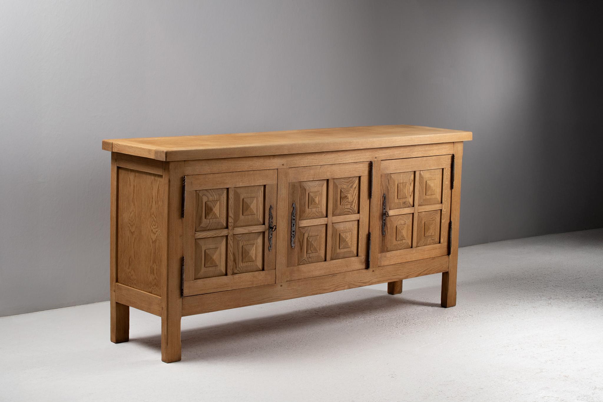 Evoking the architectural language of repetition and relief, this elongated French buffet presents a tactile meditation in solid oak. Its four-panelled façade is rhythmically punctuated by square pyramidal carvings—each block a quiet modulation of
