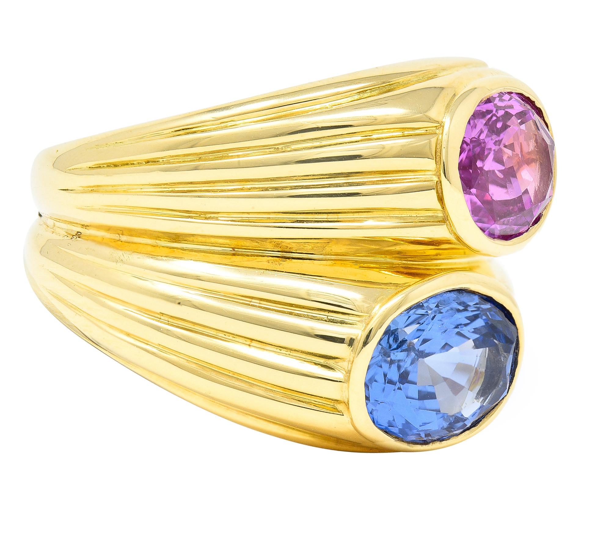 Centering two mixed cut oval sapphires one light blue and the other light purplish pink in color, Weighing 3.13 and 2.45 carats, respectively - juxtaposed North to South in high polish bezels. Flanked by fluted gold shoulders with split grooved