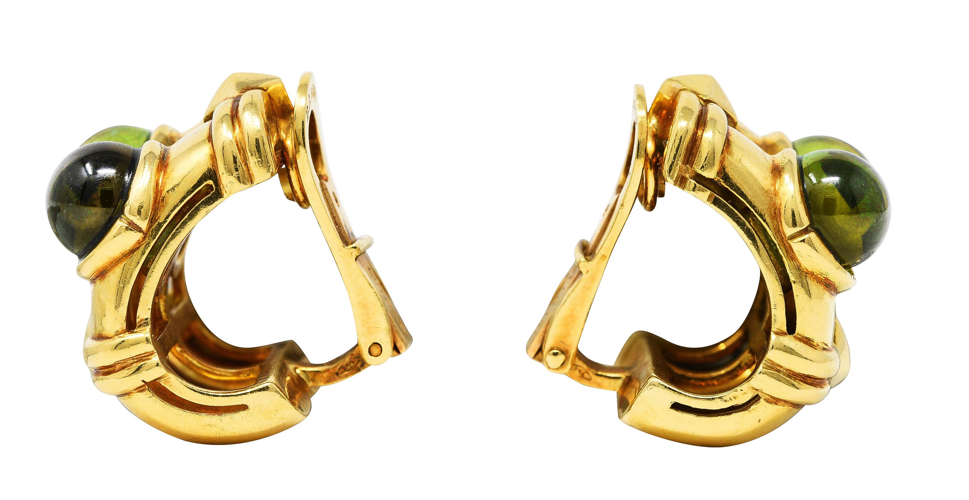 Die Ohrclip-Ohrringe sind als tief gerillte J-Ringe mit 7,0 mm großen runden Peridot-Cabochons gestaltet. Lünette gefasst und transparent mittel gelblich-grün in der Farbe. Akzentuiert durch goldene Rillenbänder und facettierte Rautenformen.