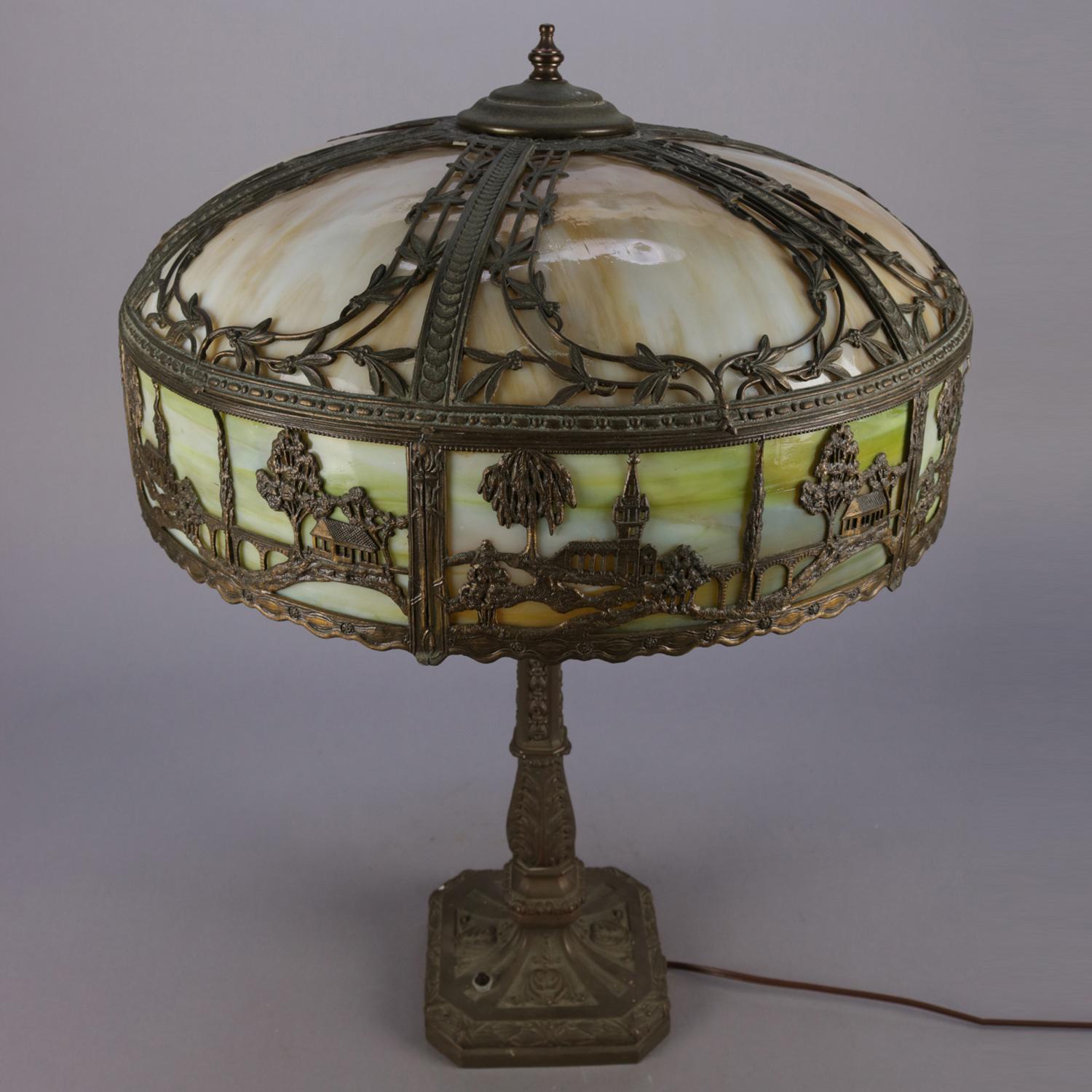 Antique Arts & Crafts Bradley & Hubbard School table lamp features scenic overlay shade with silhouette pictorial pierced frame depicting countryside scene and housing slag glass panels over independently controlled dual light cast foliate base,