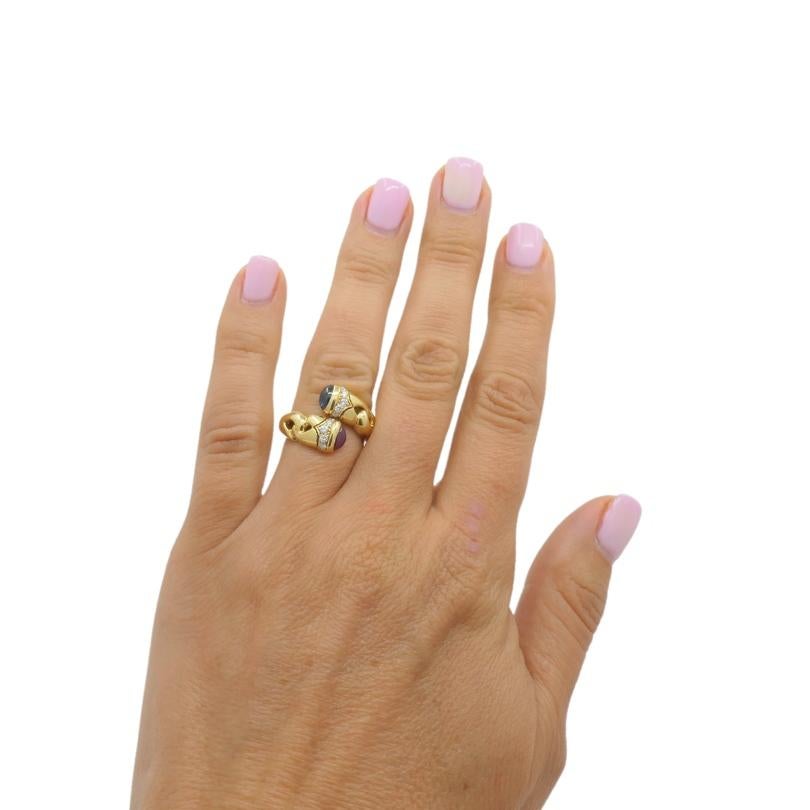 
An iconic Bulgari Doppio Cuore bypass ring features diamonds and colored gemstones. Two cabochon-cut gems—a vibrant ruby and a deep blue sapphire—gently curve towards each other. Accented with a row of sparkling diamonds, it adds just the right