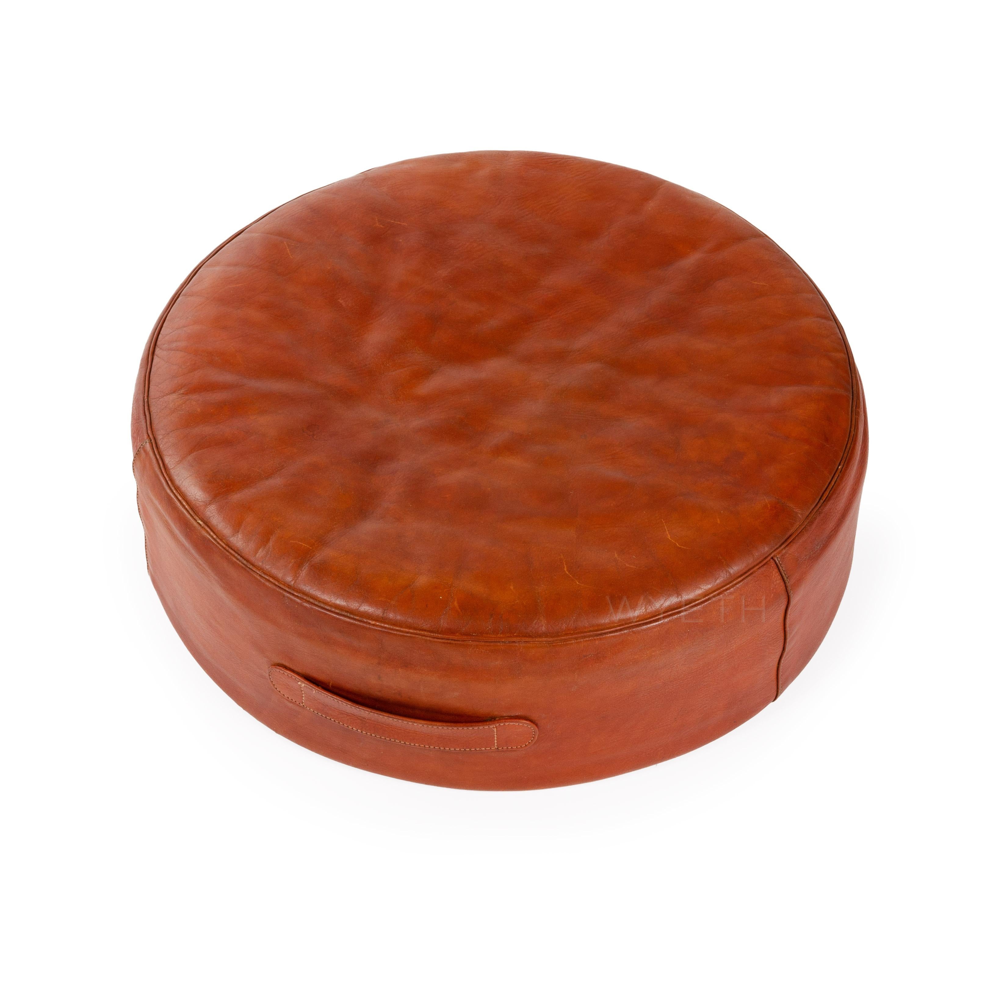 A round leather ottoman / stool retaining the original red leather upholstery and leather handle.