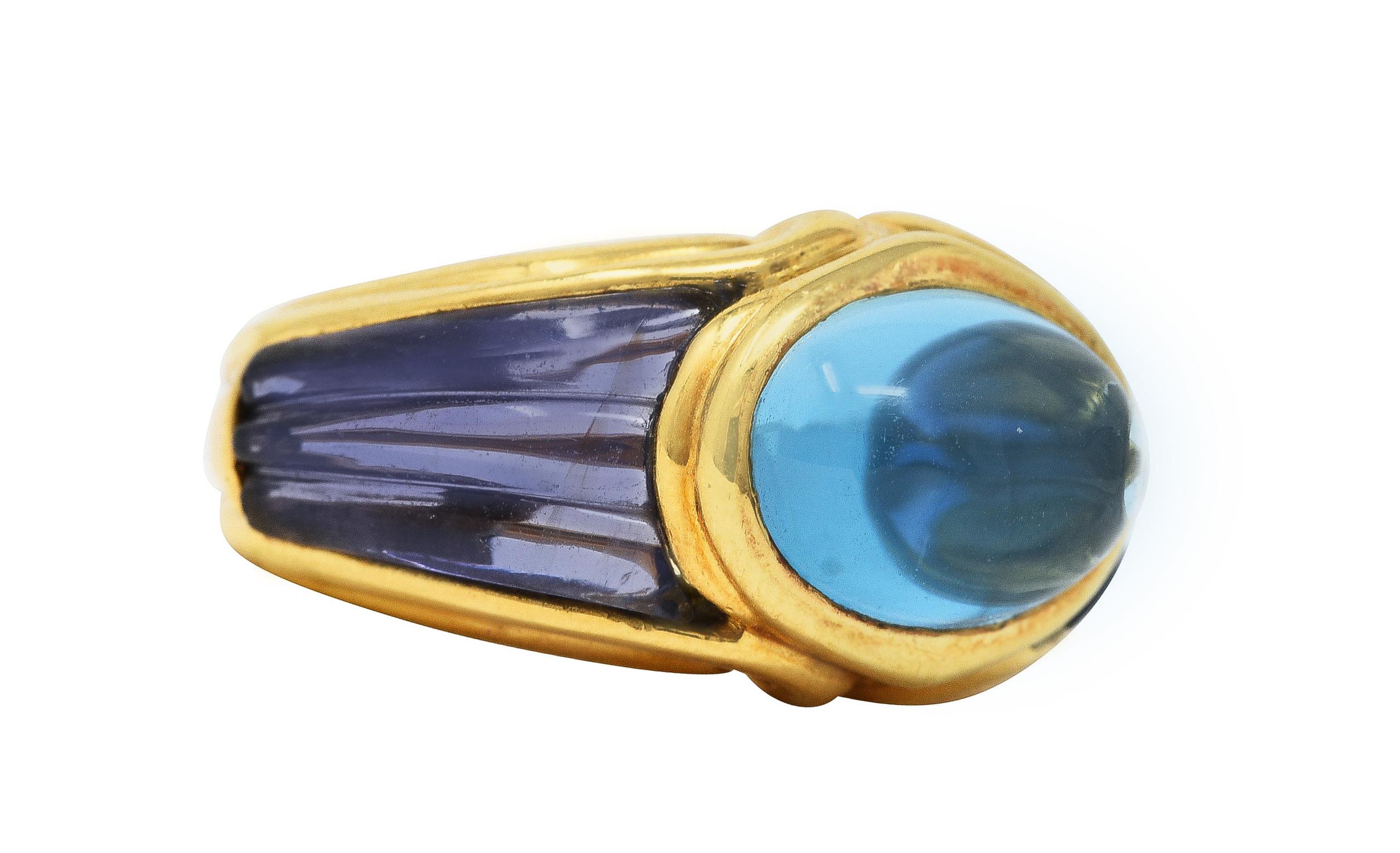 Gold band ring centers an oval cabochon blue topaz measuring approximately 10.5 x 7.5 mm. Transparent medium light sky blue. Bezel set in a polished gold surround. Shoulders are inset with carved iolite measuring approximately 11.5 x 6.5 mm. Deeply