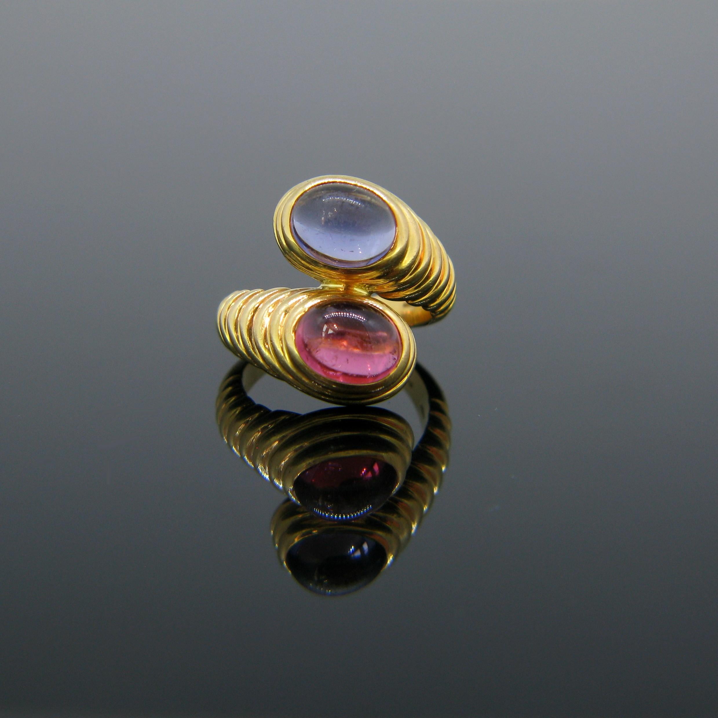 This opulent ring is fully made in 18kt yellow gold. It is set with two cabochon cut stones: an iolite and a pink tourmaline. They are arranged in a bypass/crossover design. The band is textured with round ridges. It is signed Bulgari inside the