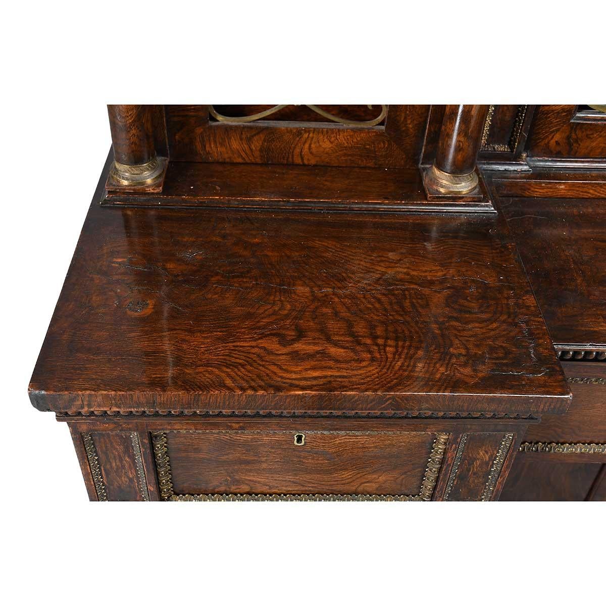 A Regency period bookcase in figured elm and grain-painted ‘elm’ and adorned with brass accents. The fall-front at the center opens to reveal a writing surface, drawers, and compartments. Attributed to George Bullock. The brass grillwork on the